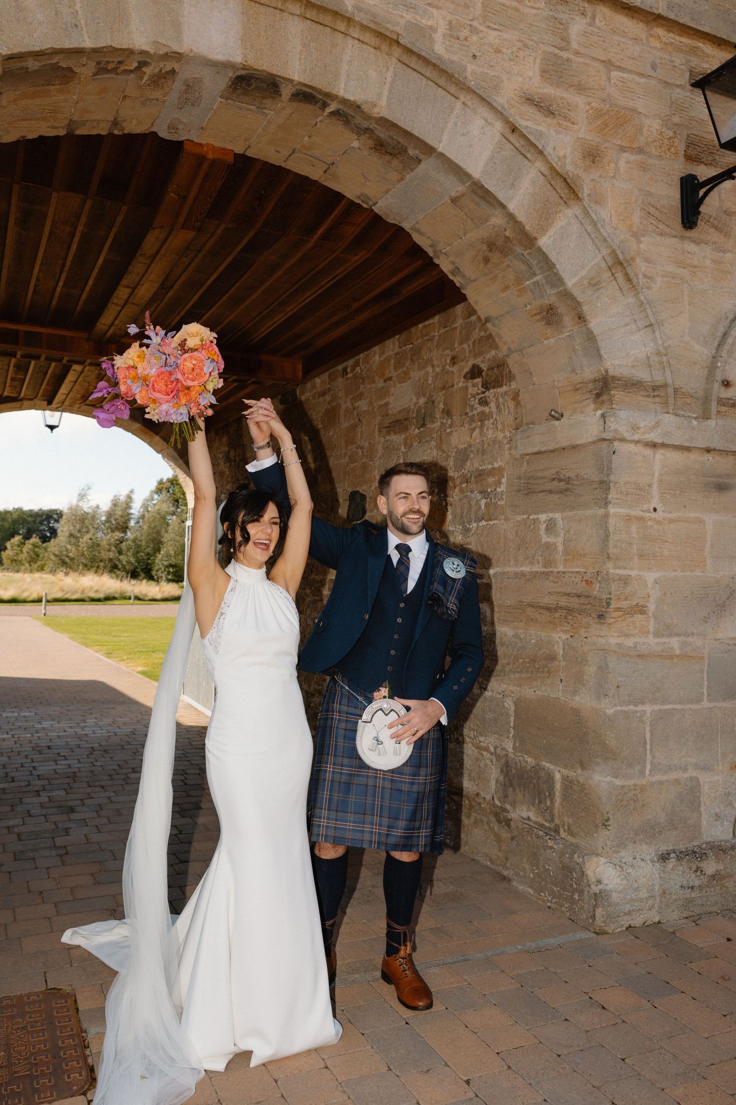 Bride in white gown and groom in blue jacket and tartan kilt celebrate under stone arch, raising colorful bouquet.