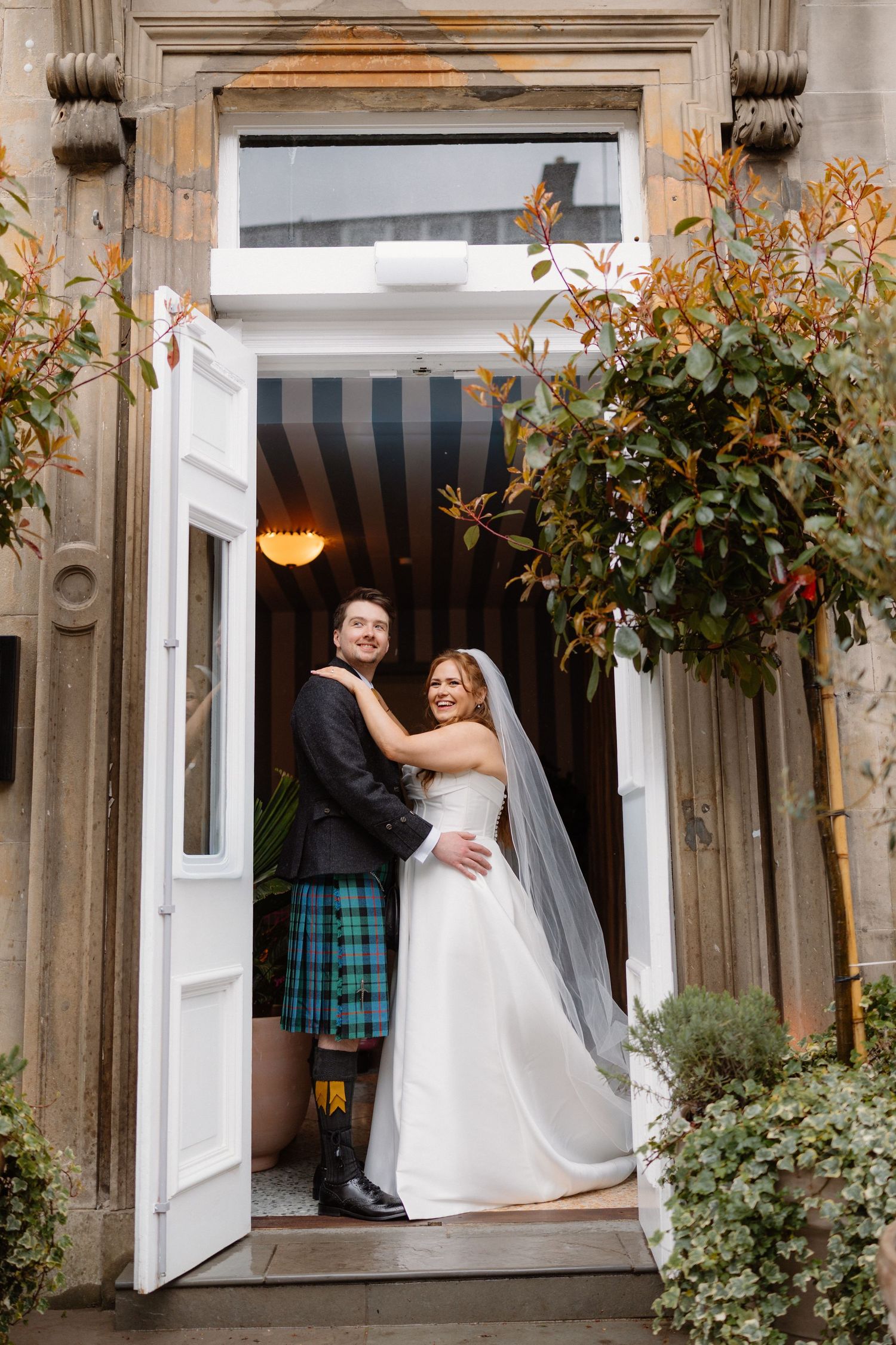 Bride and groom in Scottish kilt posing together in doorway of The Hoxton Edinburgh on wedding day.