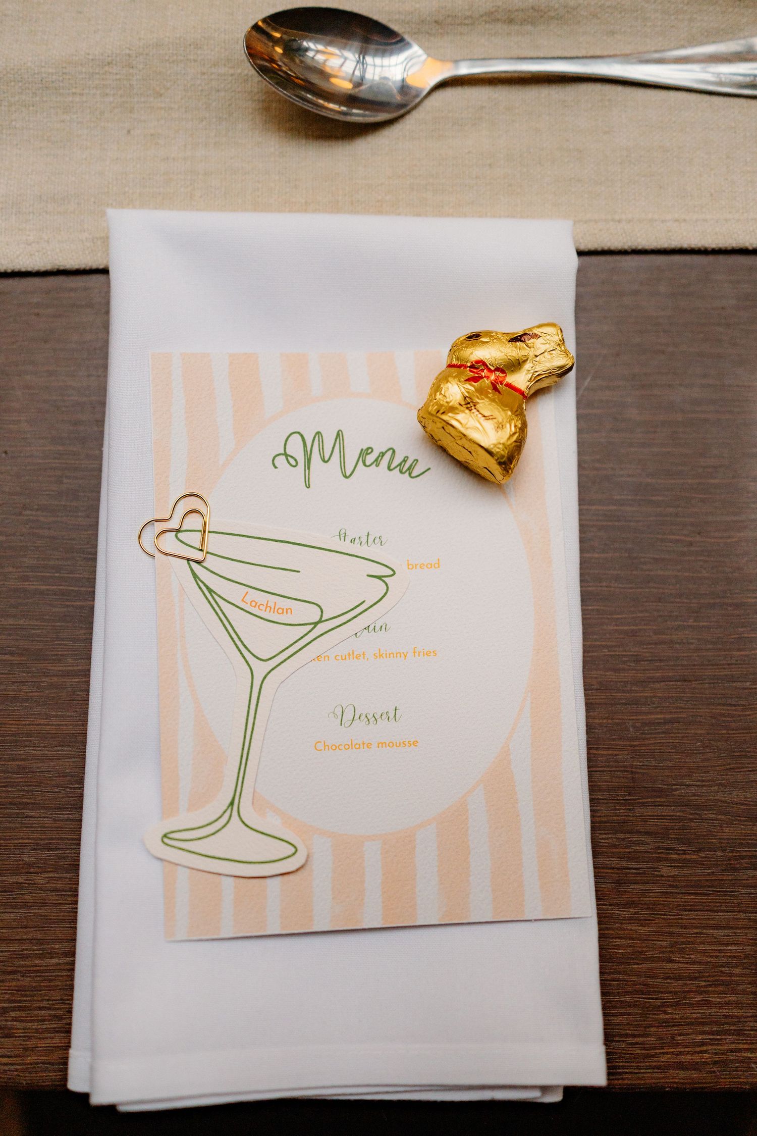 Elegant wedding menu card with martini glass illustration and gold foil chocolate bunny on a white linen napkin.