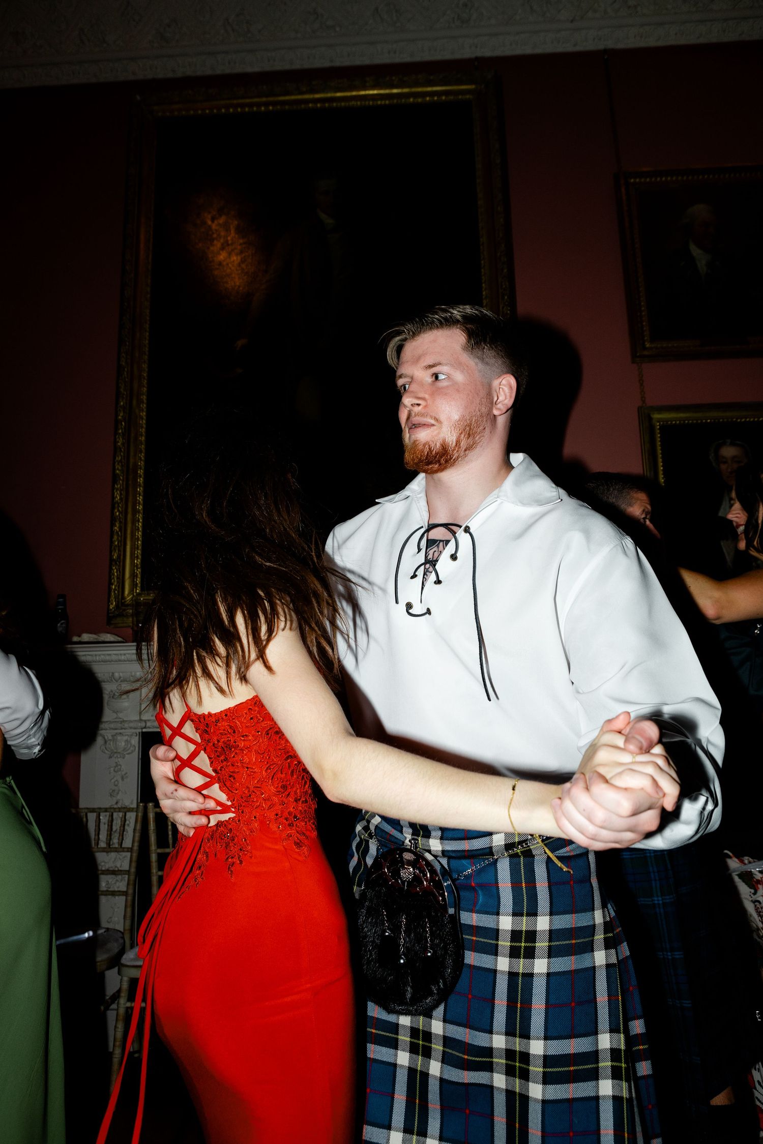 Couple dancing at wedding reception, man in white shirt and tartan kilt, woman in red dress.