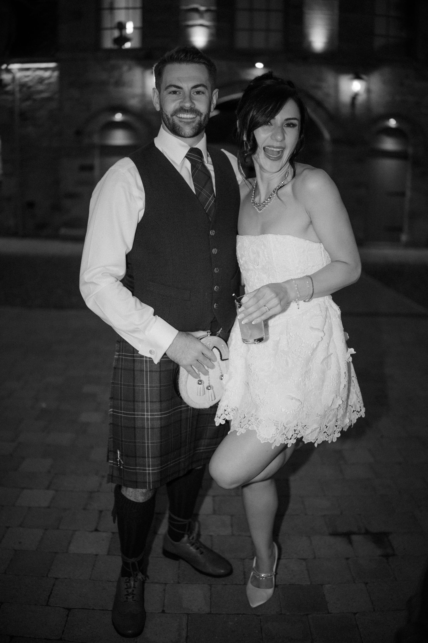 Black and white photo of a groom in a kilt and bride in a short white lace dress posing at a wedding.