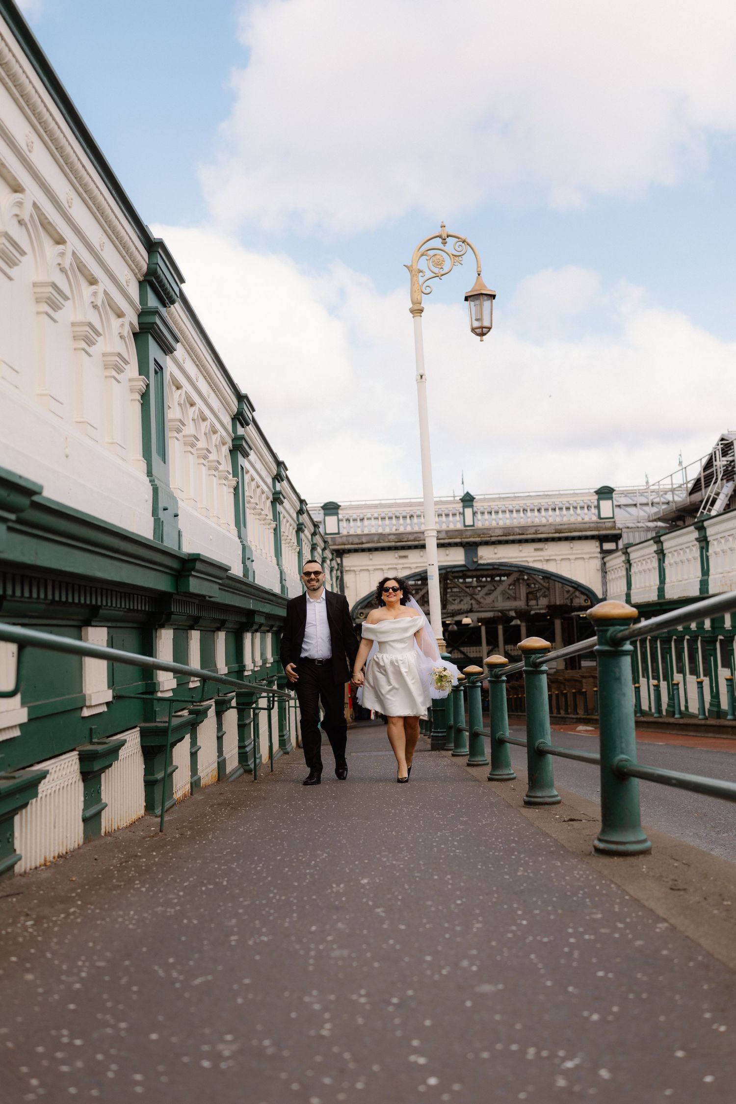 Couple walking hand in hand along a Victorian pier with green railings and white ornate architecture.