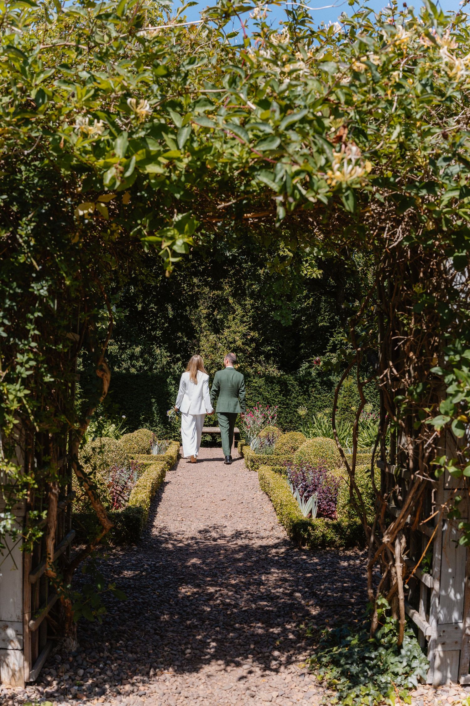 Newlywed couple walking hand-in-hand through a lush green garden archway on their wedding day.