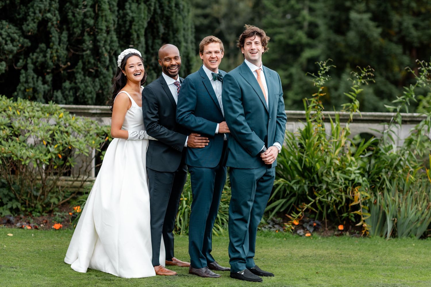 Wedding party members share candid moments in garden with evergreen trees.