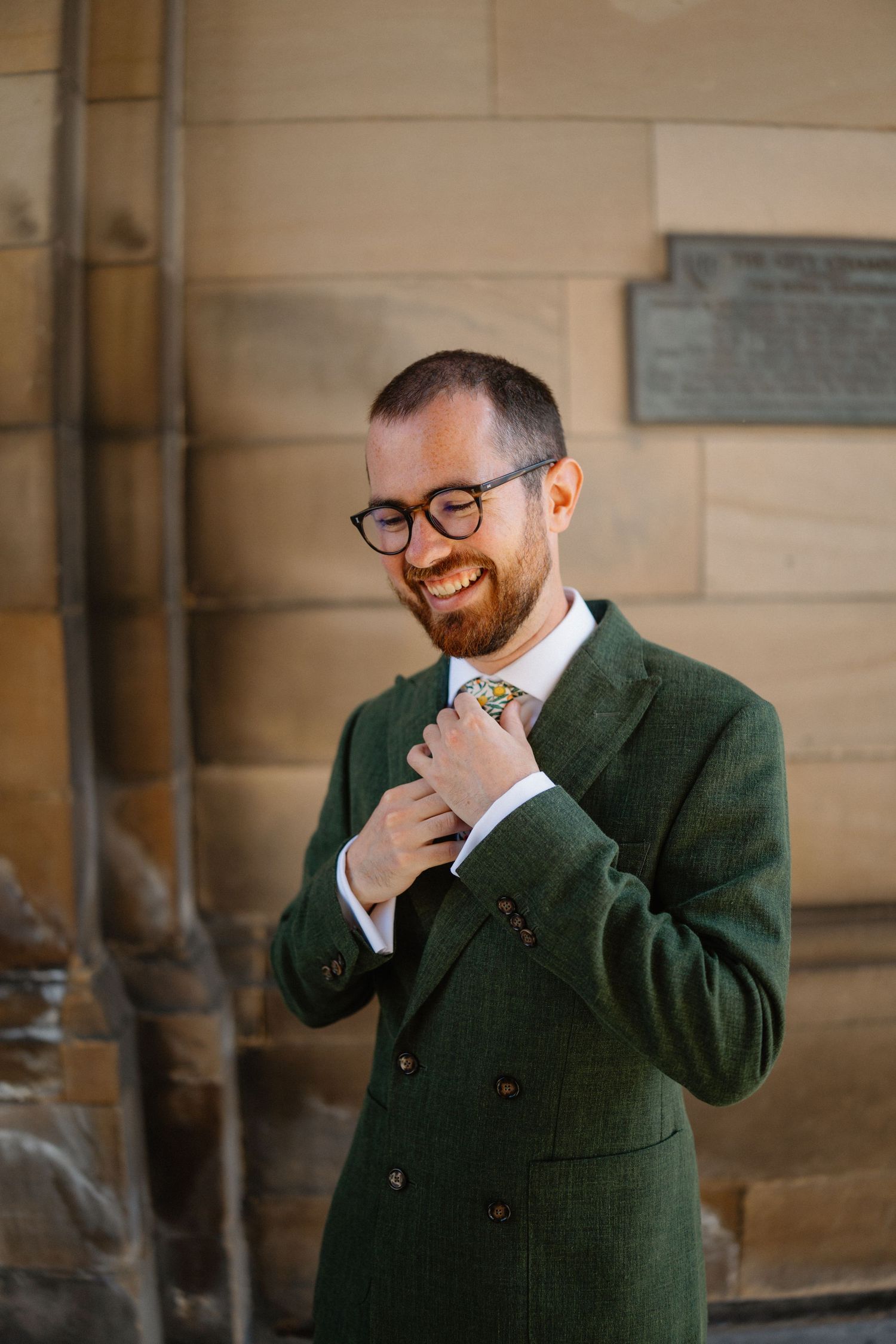 Person in dark green suit jacket and glasses adjusting their tie.