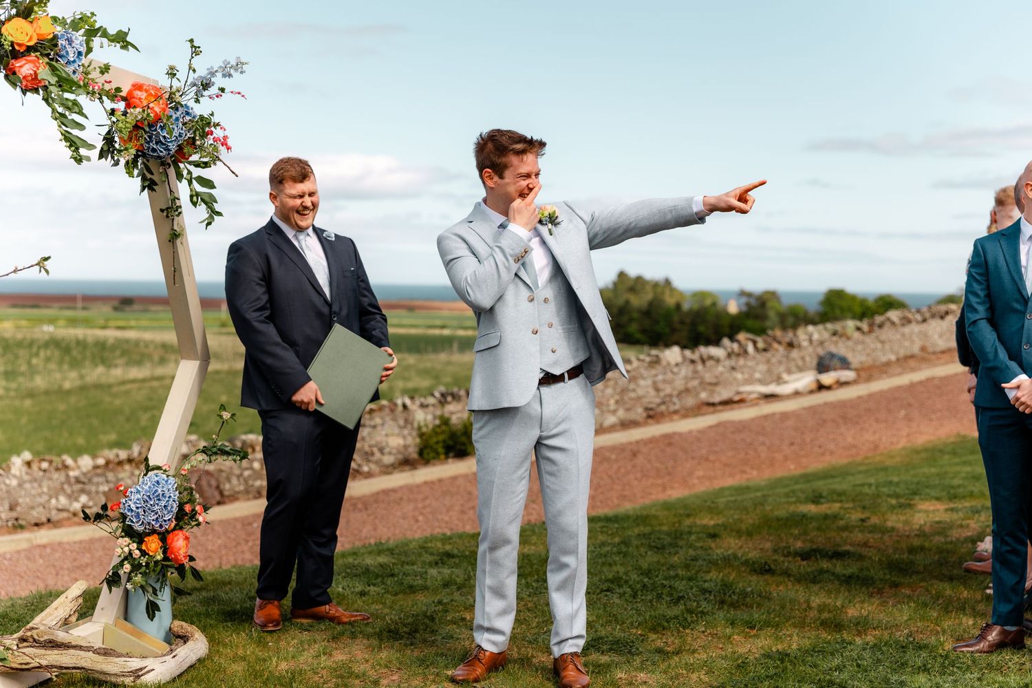 Person in light gray suit gestures while standing outdoors with floral decorations.