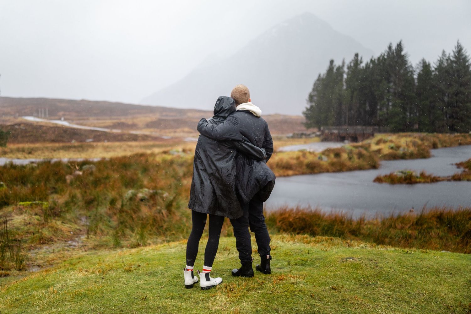 Two people embracing while standing on a hill overlooking a misty Scottish landscape.