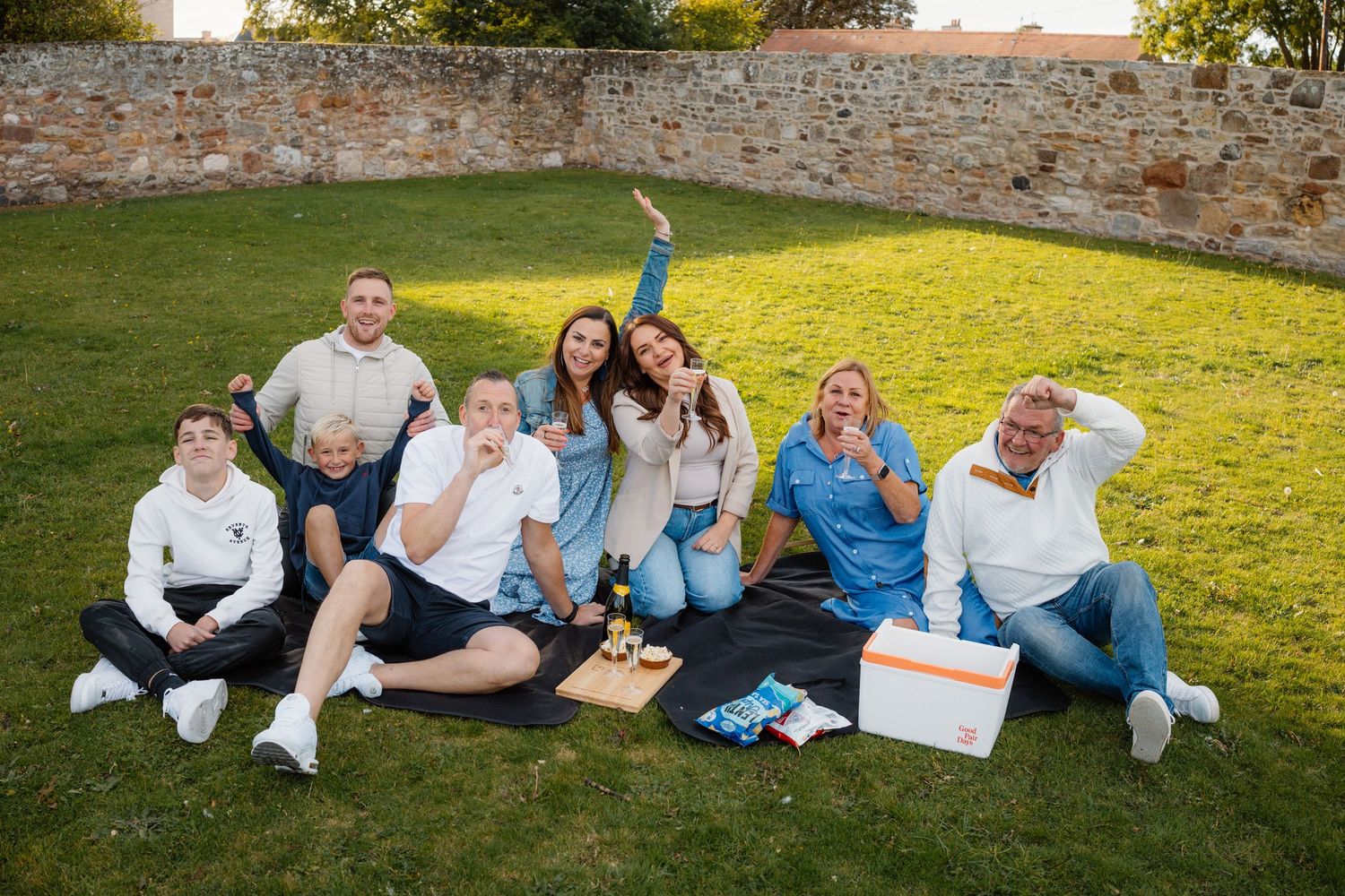 Group of friends and family having picnic on sunny lawn near stone wall.