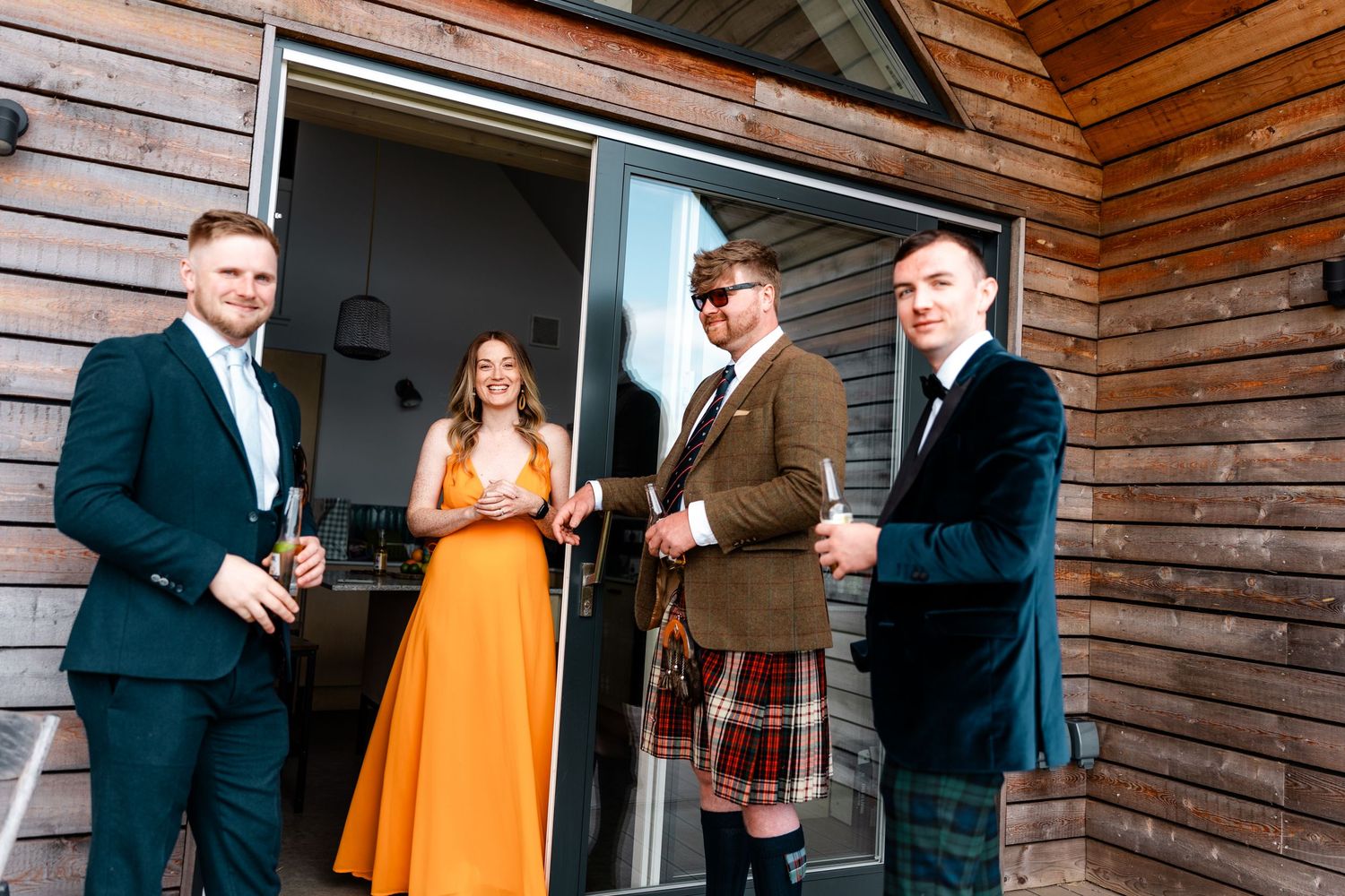 Wedding party members in formal attire and kilts gathered outside a rustic wooden building.