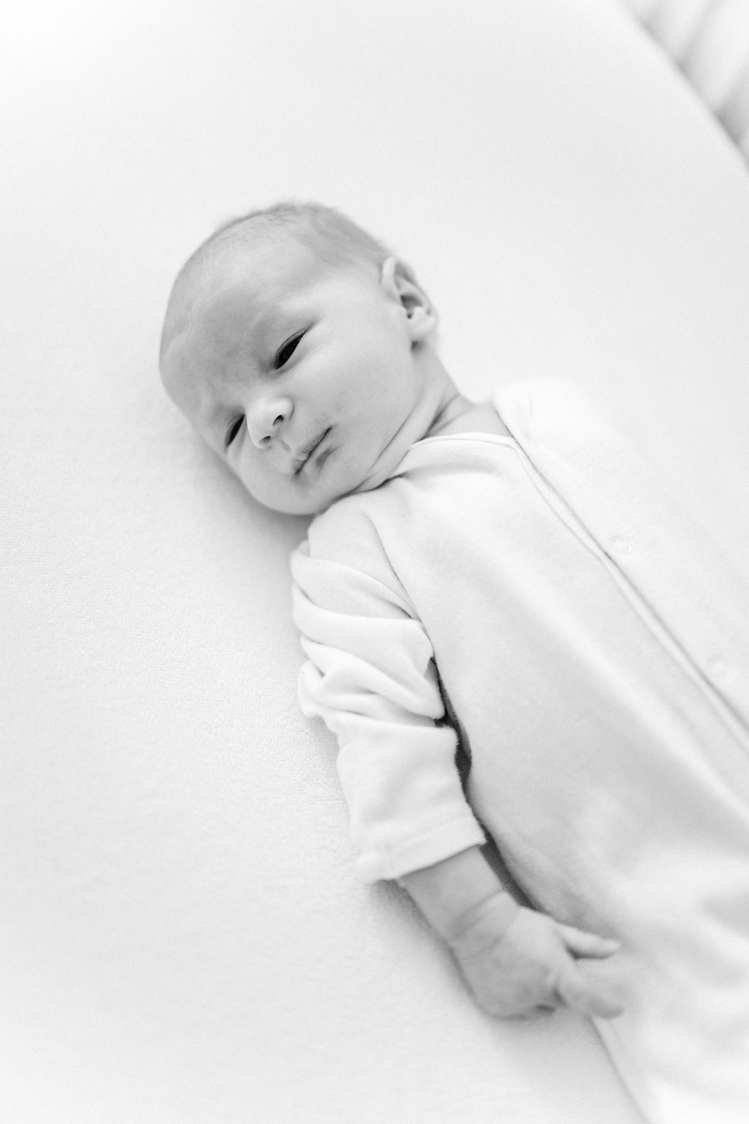 A newborn baby wearing white clothing rests peacefully on soft white bedding.