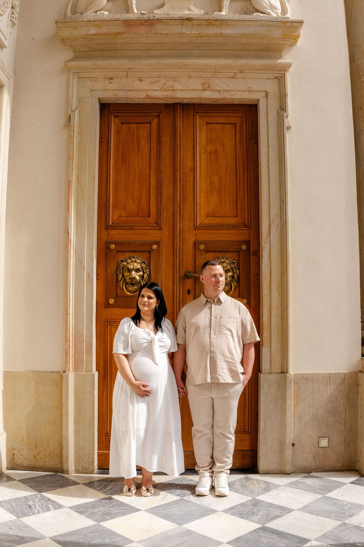 A couple poses against ornate wooden doors and white stone walls in a classic architectural setting.