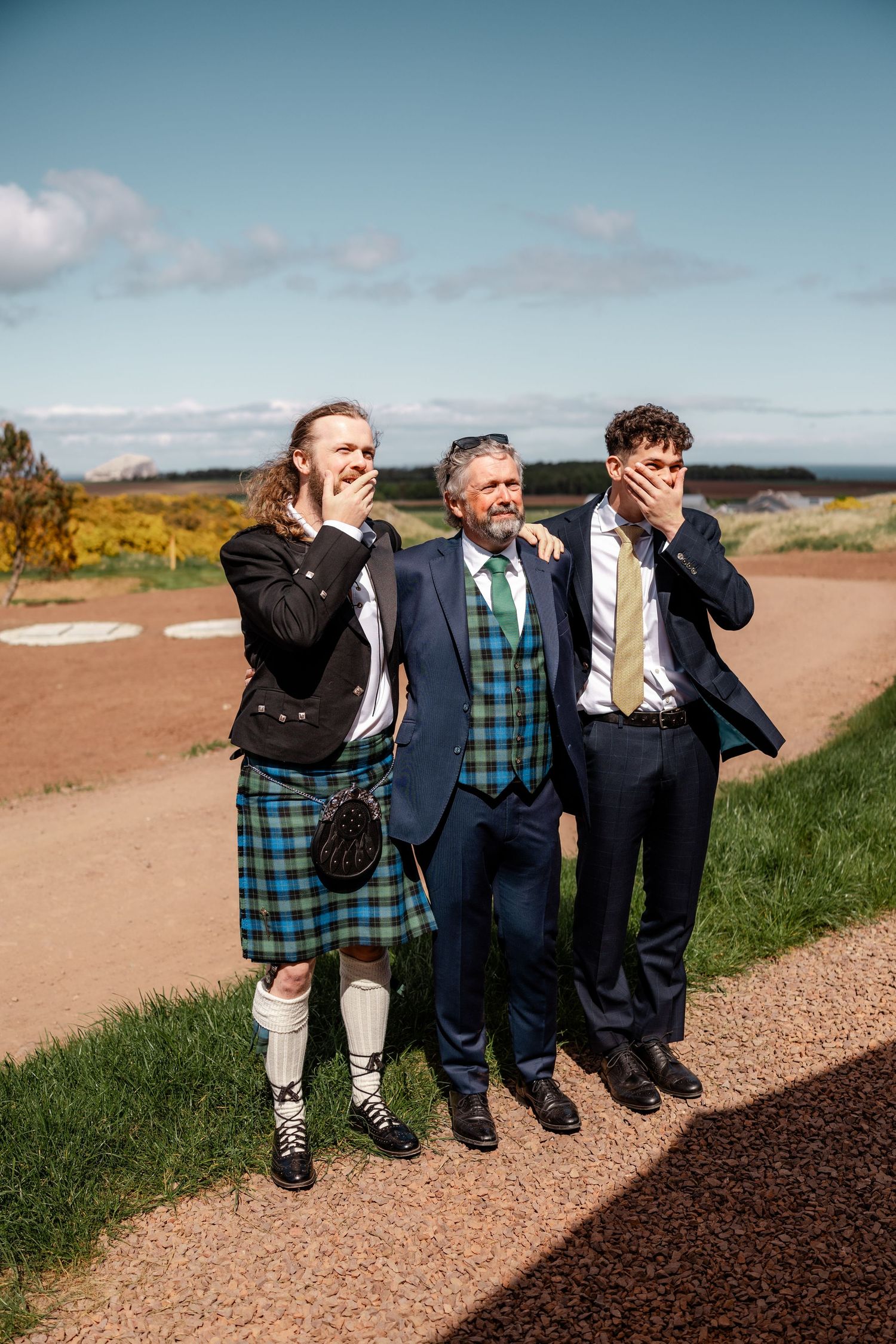 Three men in formal wedding attire laugh outdoors, one wearing a traditional Scottish kilt on a sunny day.