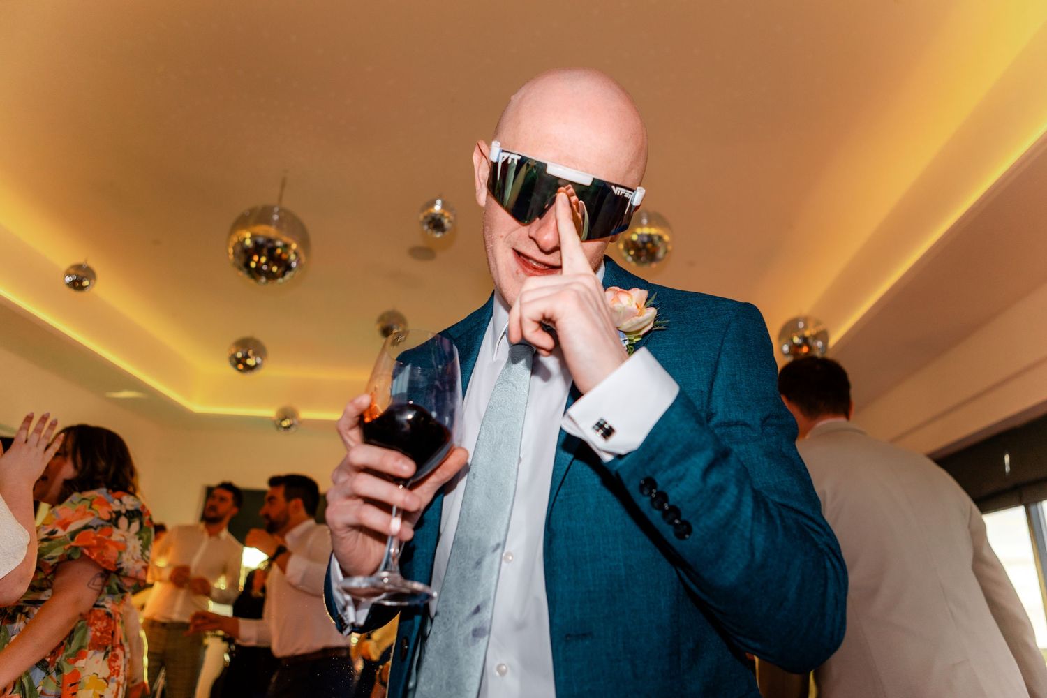 Bald man in teal suit wearing oversized sunglasses holding red wine at wedding reception.