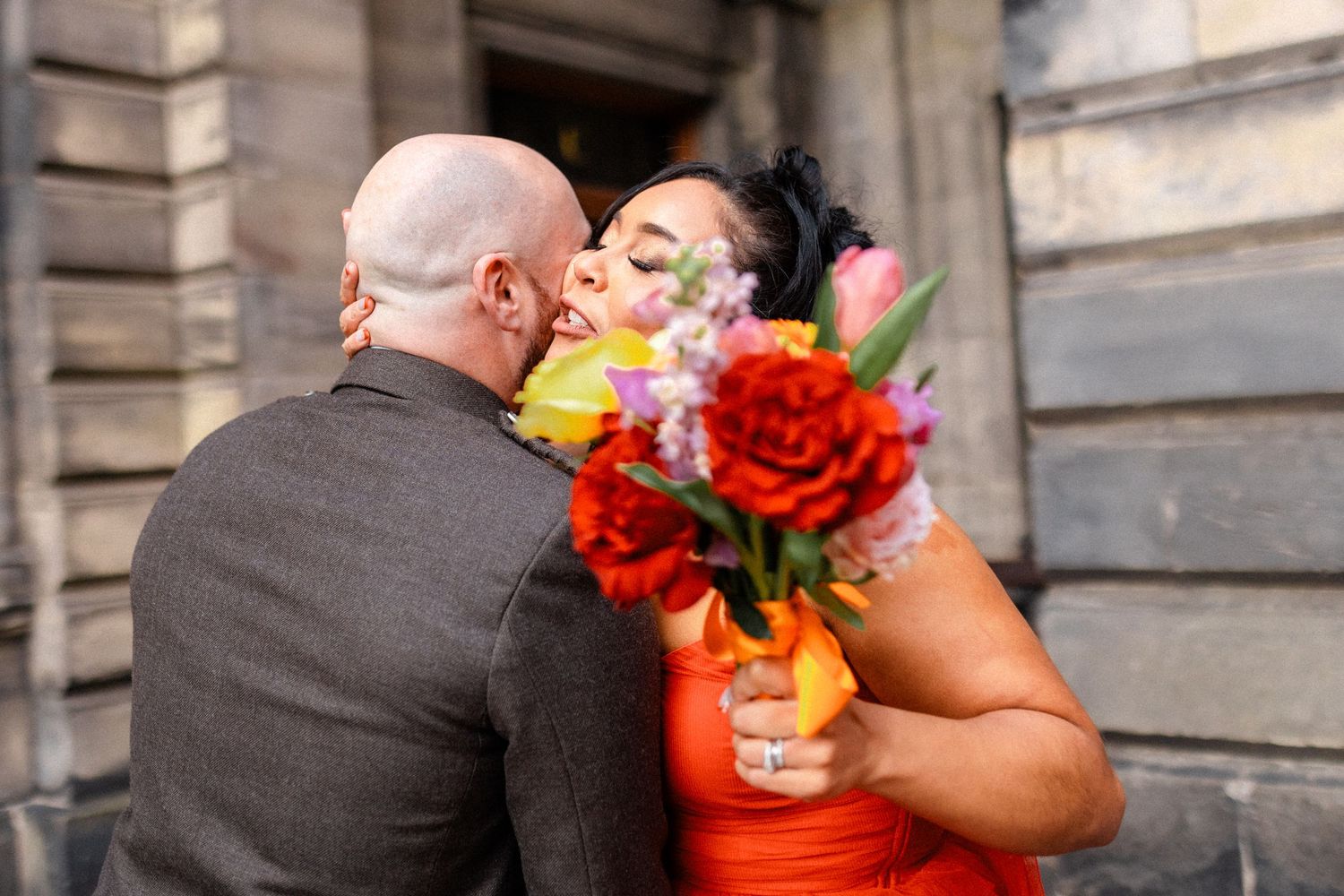 Bride in orange dress kissing bald groom while holding vibrant bouquet of colorful flowers outdoors.