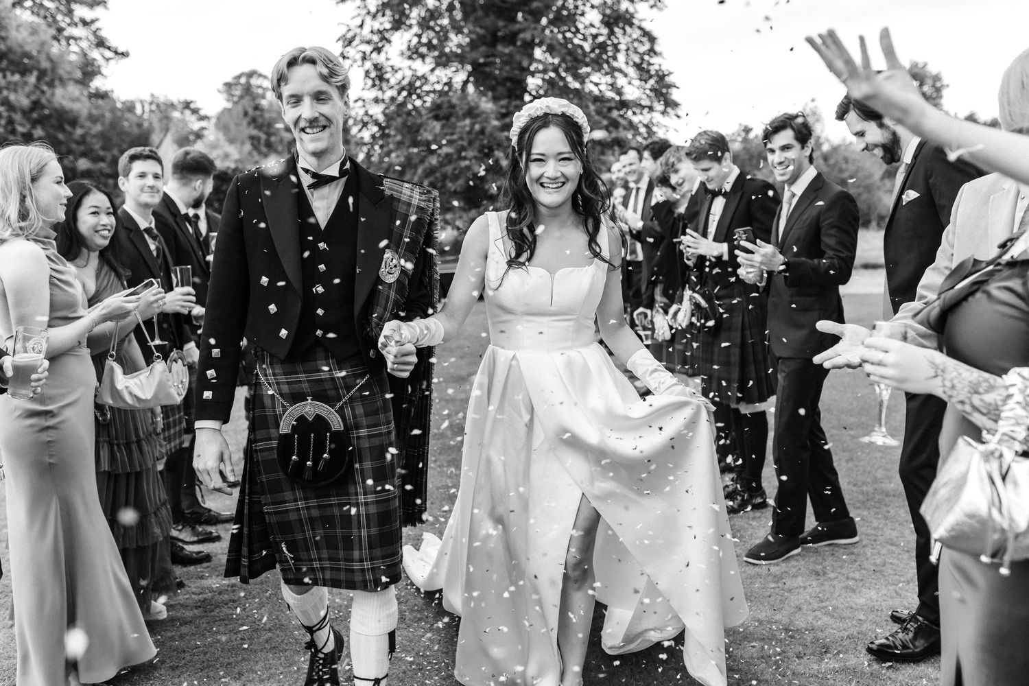 Bride and kilted groom walk through confetti shower surrounded by cheering wedding guests outdoors.