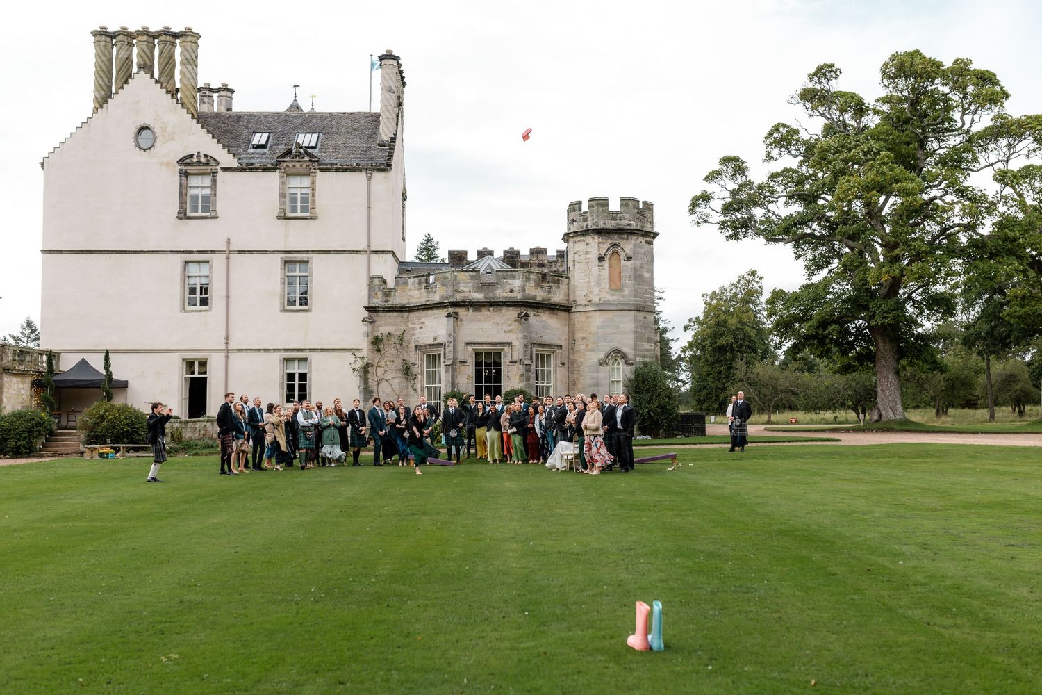 Wedding guests gather on the lawn of a historic castle estate for a group photo on a cloudy day.