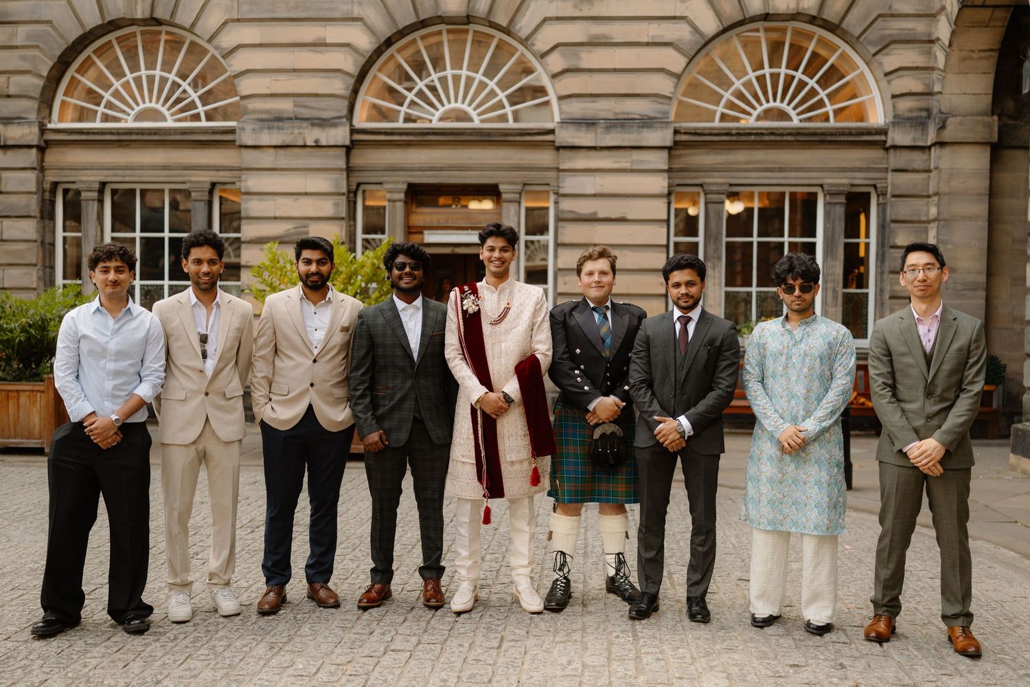 Group of nine men in formal and traditional attire posing outside a historic stone building.