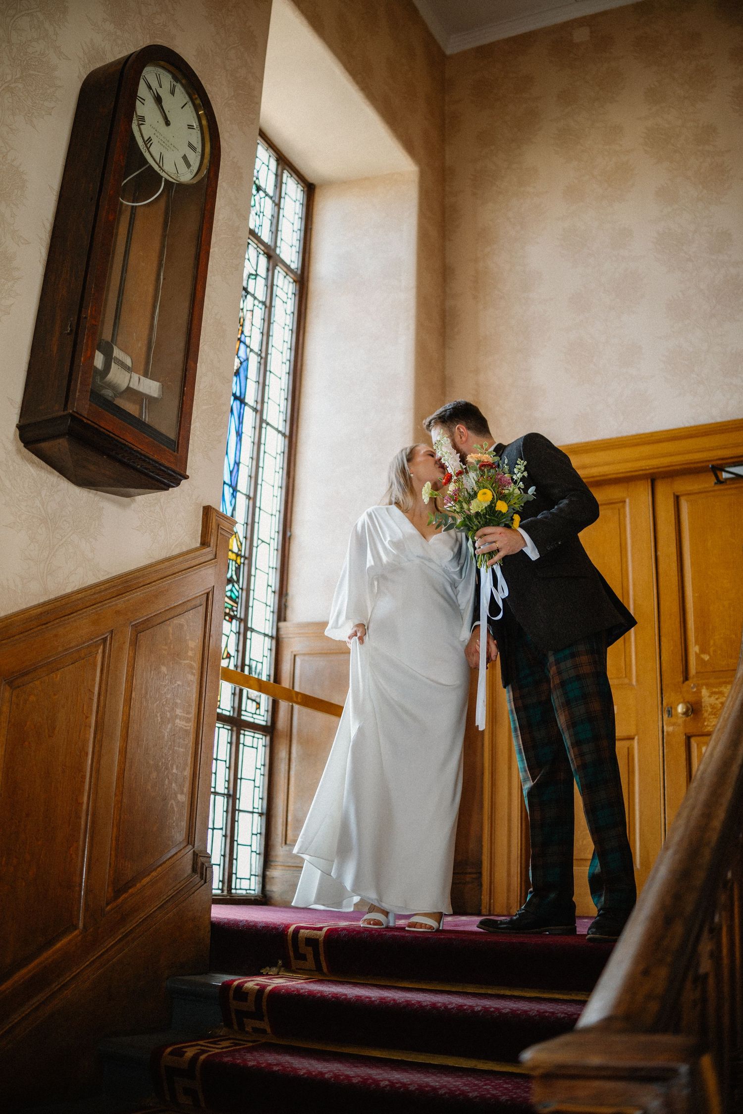 Bride and groom share a kiss on carpeted stairs beside a vintage clock and stained glass.