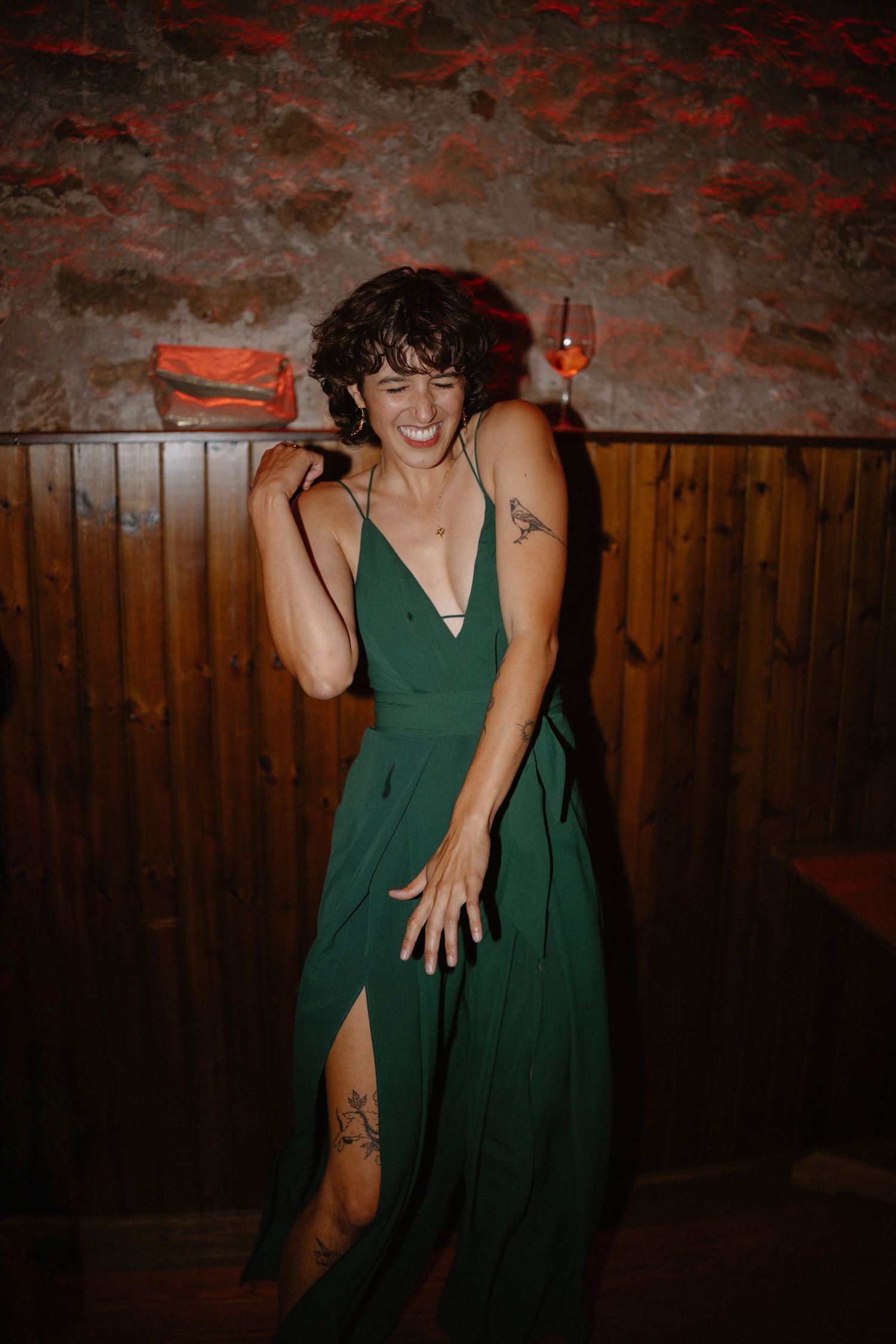 Woman in emerald green slit dress laughing and dancing in a dimly lit rustic stone venue.