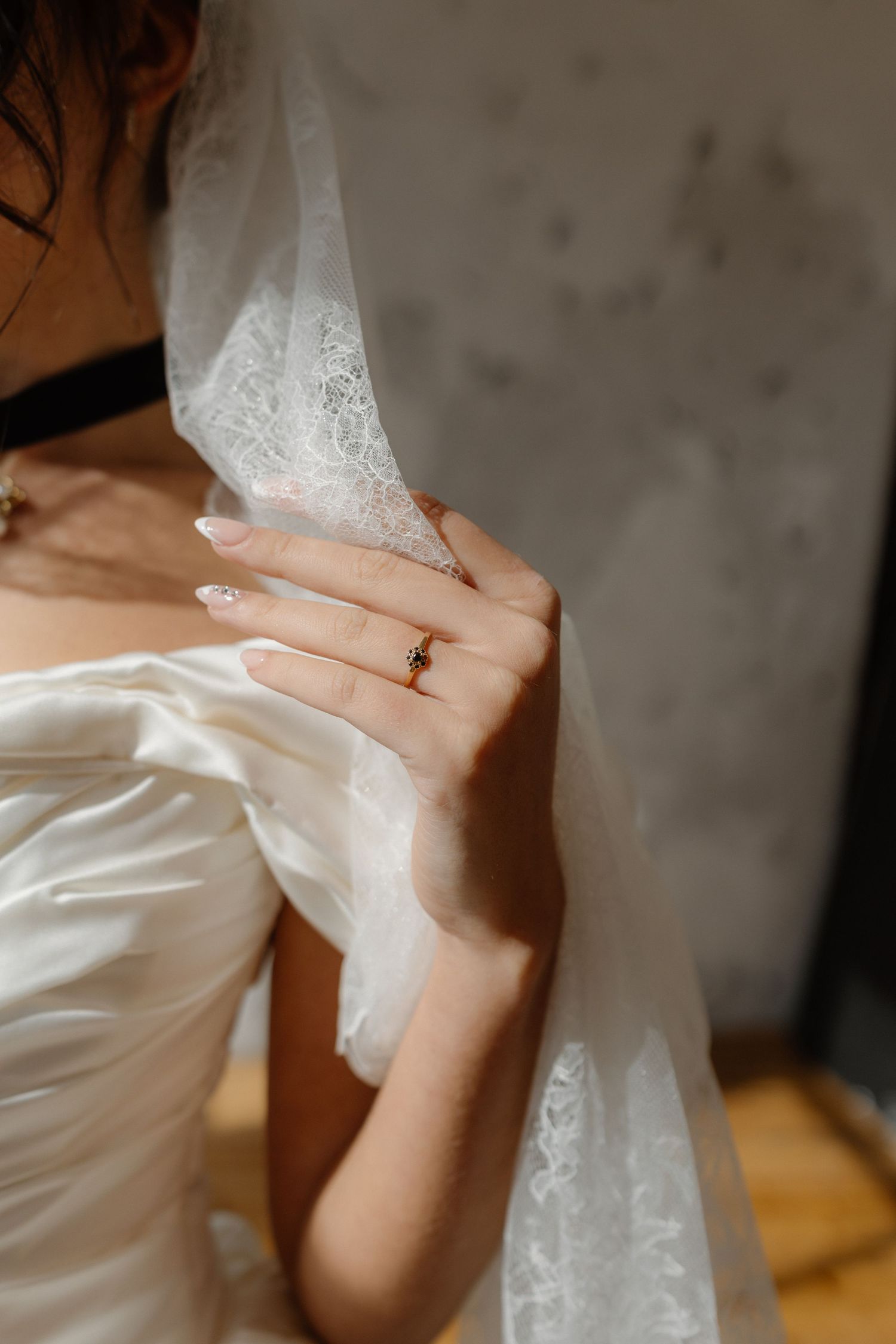 Close-up of bride's hand holding lace veil, wearing gold ring with red gemstone, white satin dress.