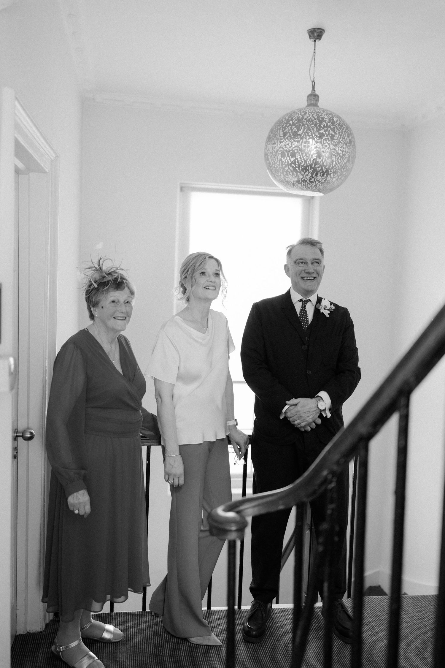 Black and white photo of bride with parents smiling on staircase under decorative chandelier at wedding.