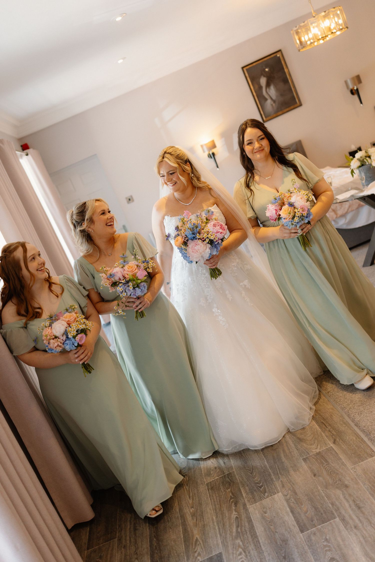 Bride in white lace gown with three bridesmaids in sage green dresses holding pastel bouquets.
