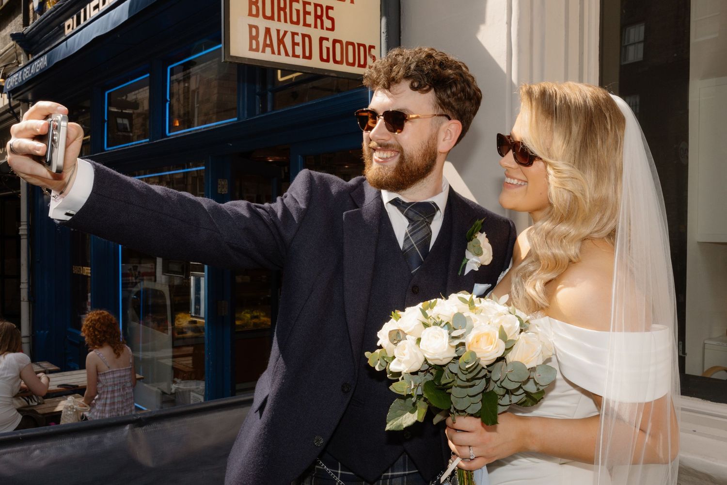 Newlywed couple taking a selfie outside a burger shop, bride holding white bouquet in wedding dress and groom in suit.
