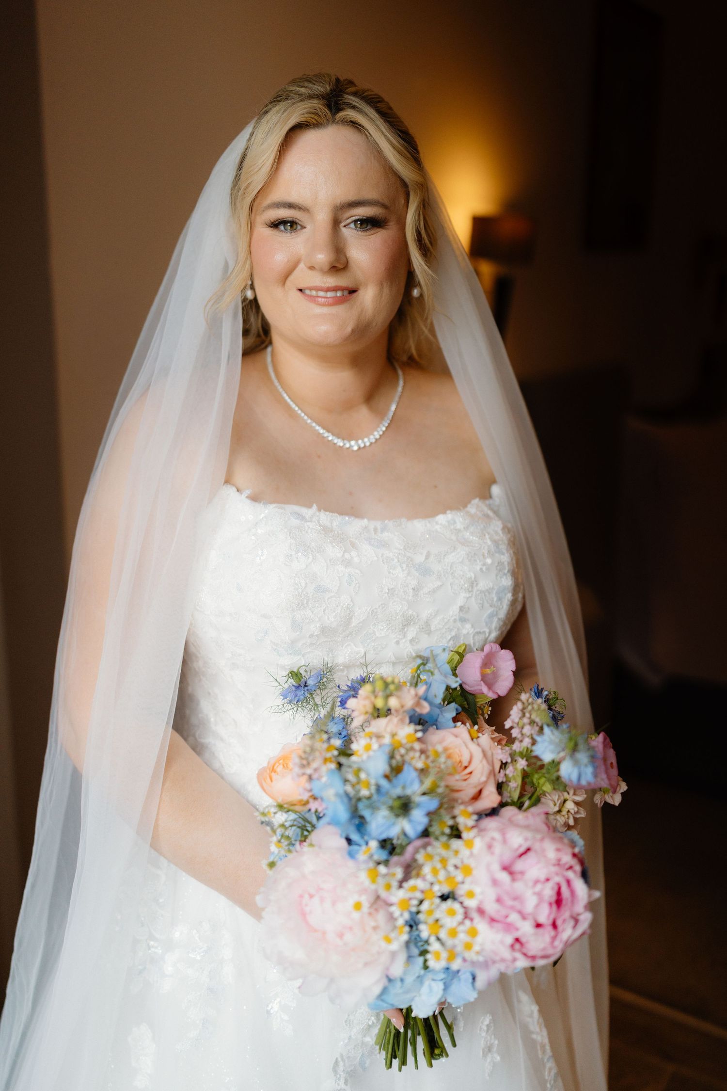 Bride in white lace gown and veil holds pastel bouquet of pink peonies and blue flowers, smiling indoors.