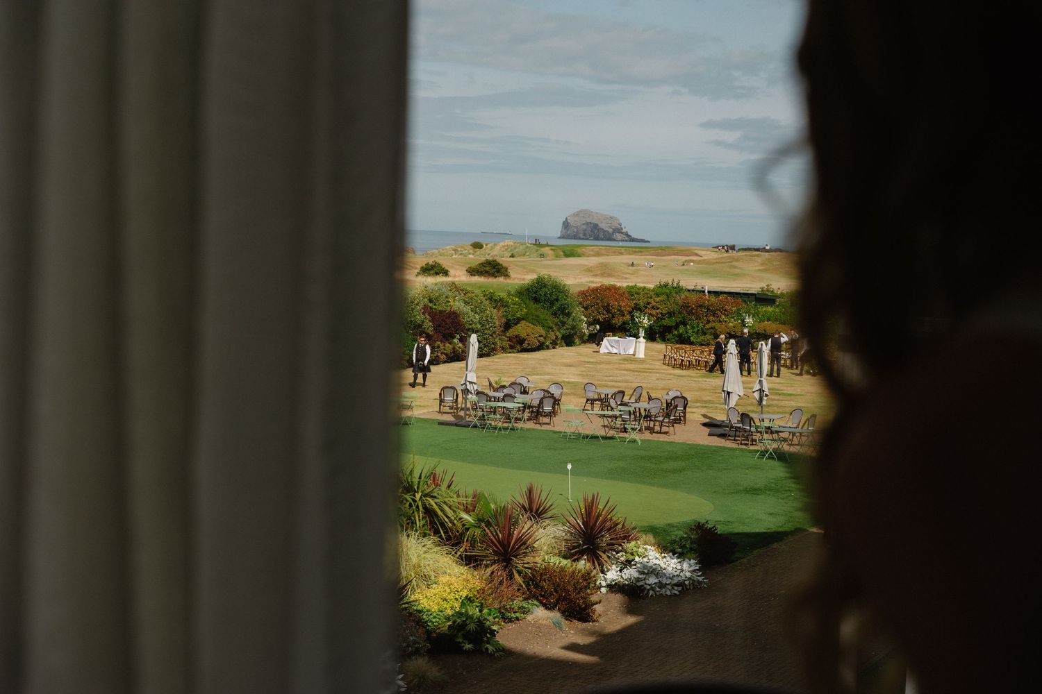 View through a window of a lush golf course with garden sculptures and a rocky coastal hill under a blue sky.
