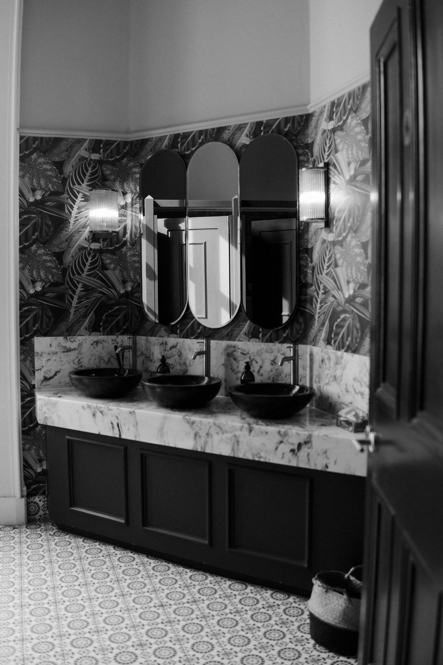 Modern bathroom with black wainscoting, patterned wallpaper, and geometric tile flooring illuminated by wall sconces.