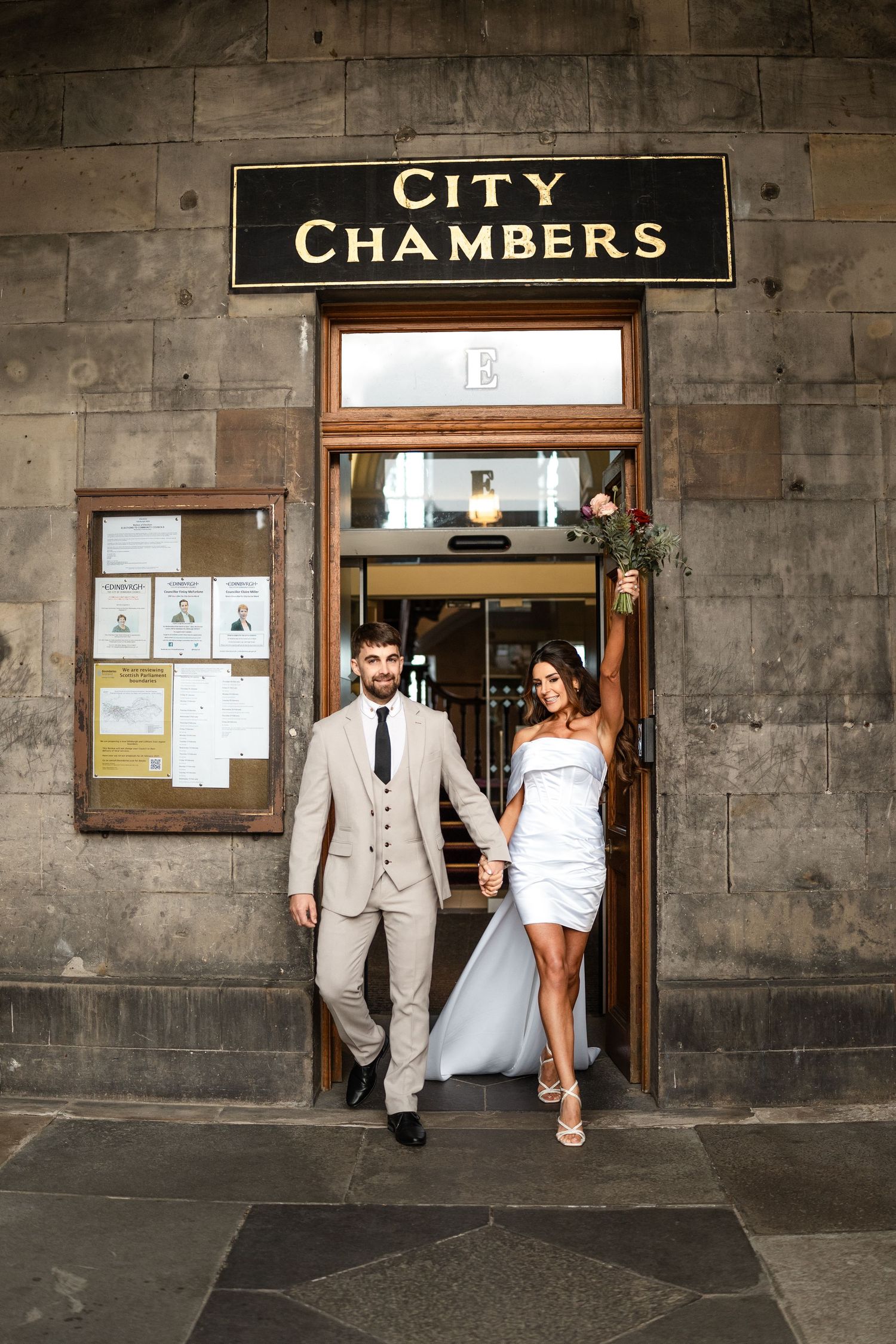 Wedding couple exits City Chambers building with stone facade.