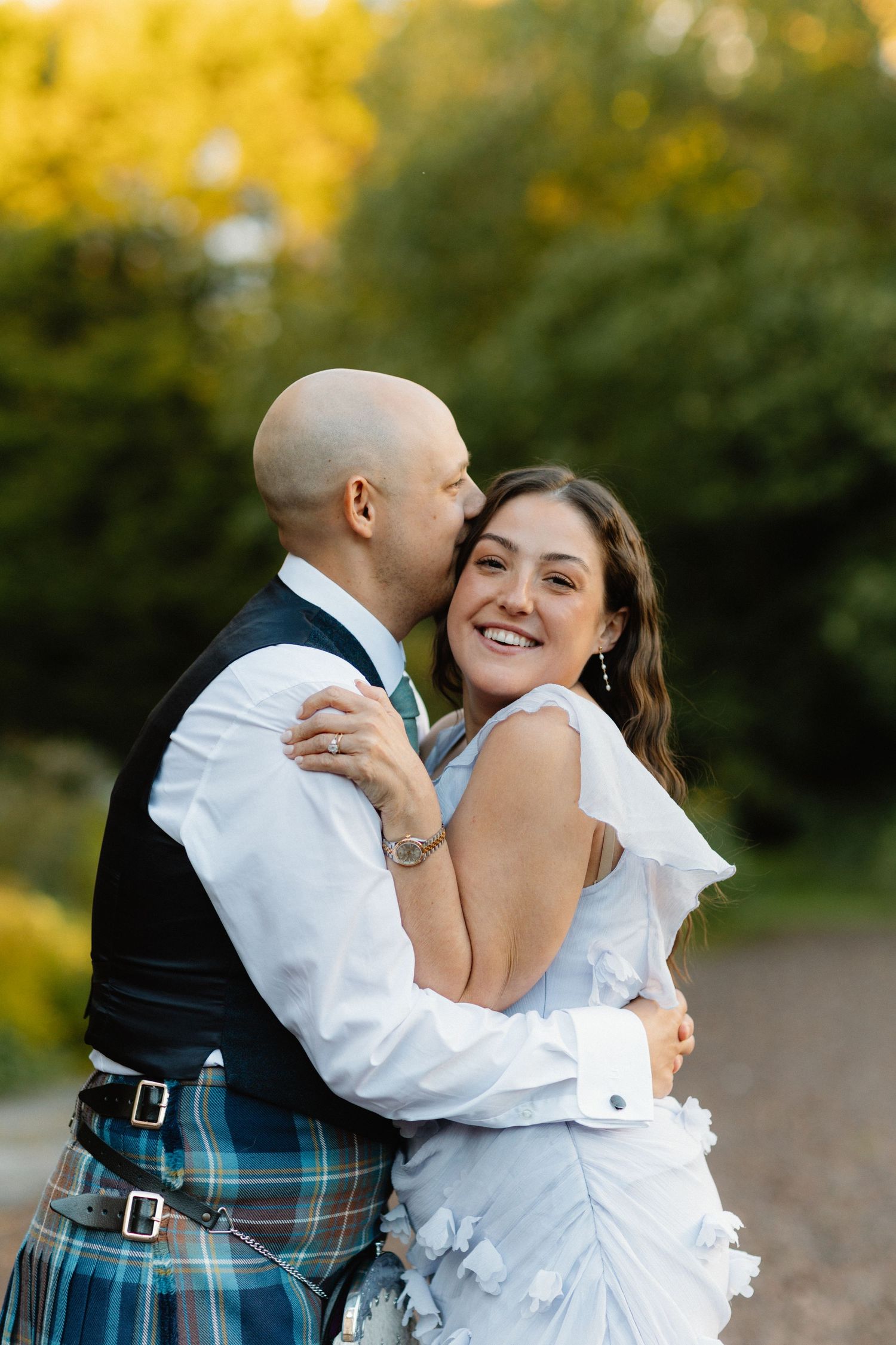 Couple in wedding attire embracing outdoors with autumn foliage in background.