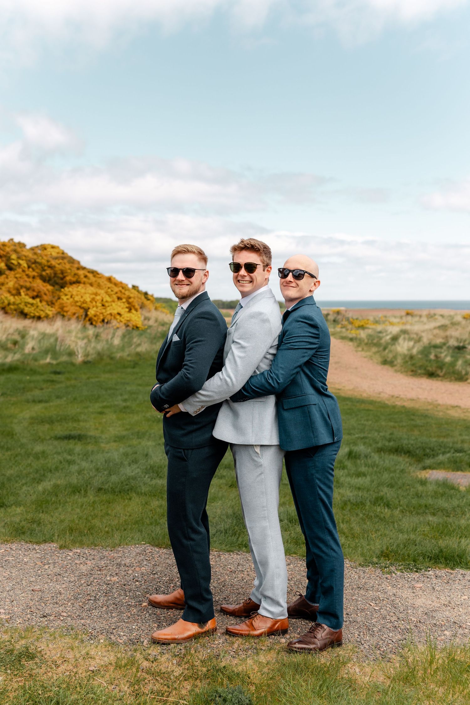 Three groomsmen in suits pose playfully on a coastal path at sunset.