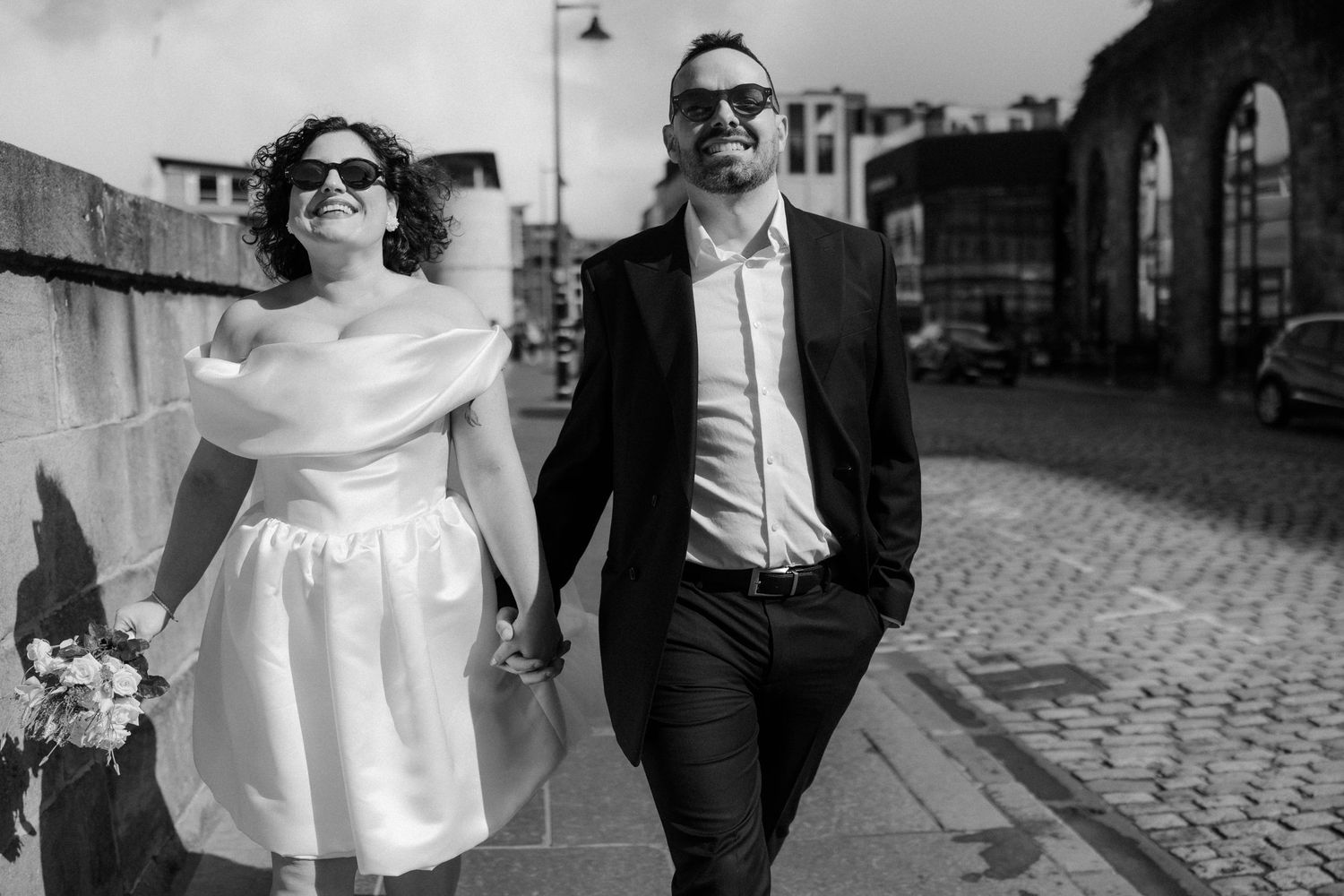 Black and white photo of a smiling bride and groom in sunglasses walking hand in hand on a cobblestone street.