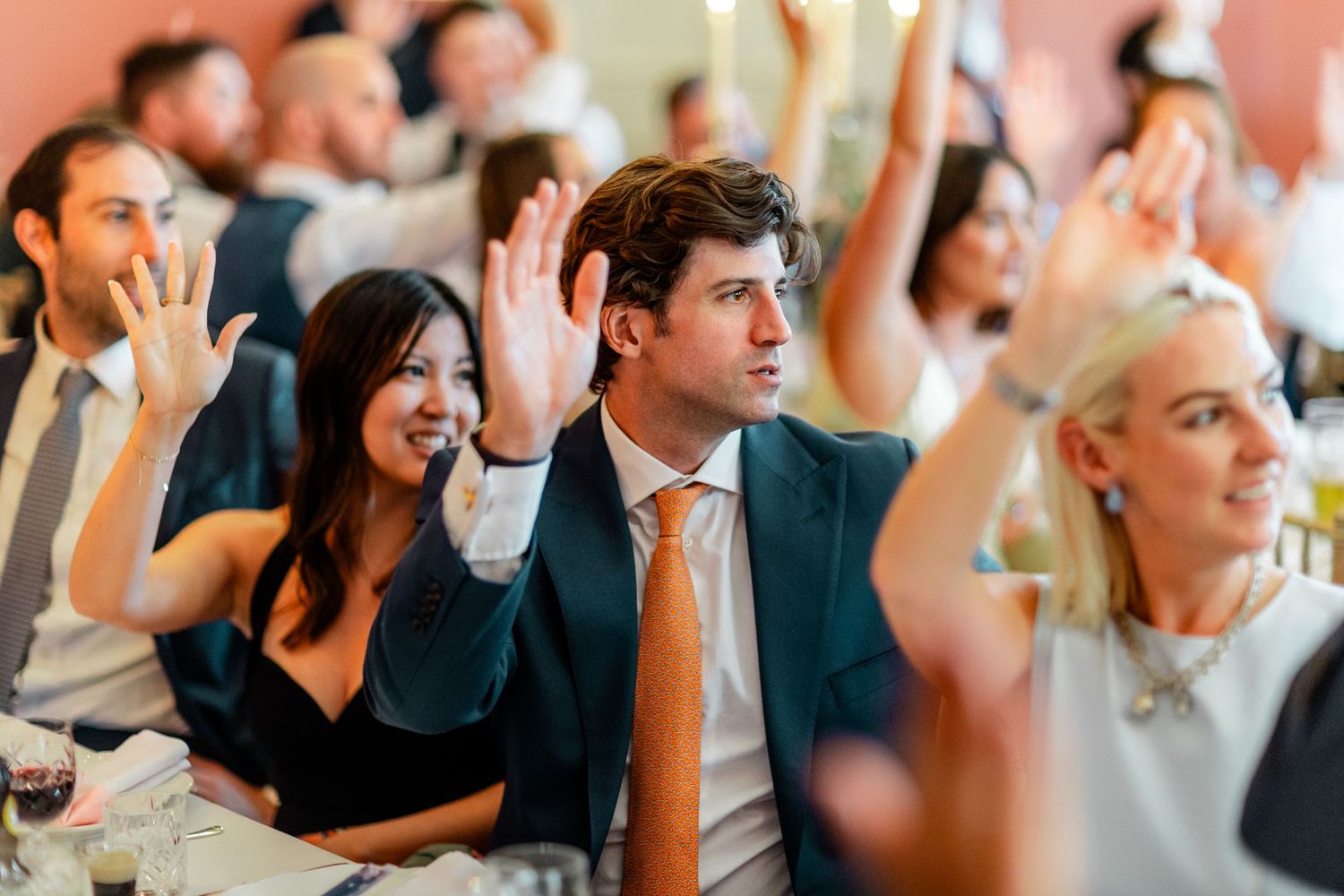 Wedding guests raise hands at a reception dinner inside an elegant venue, smiling and engaged during a fun activity.