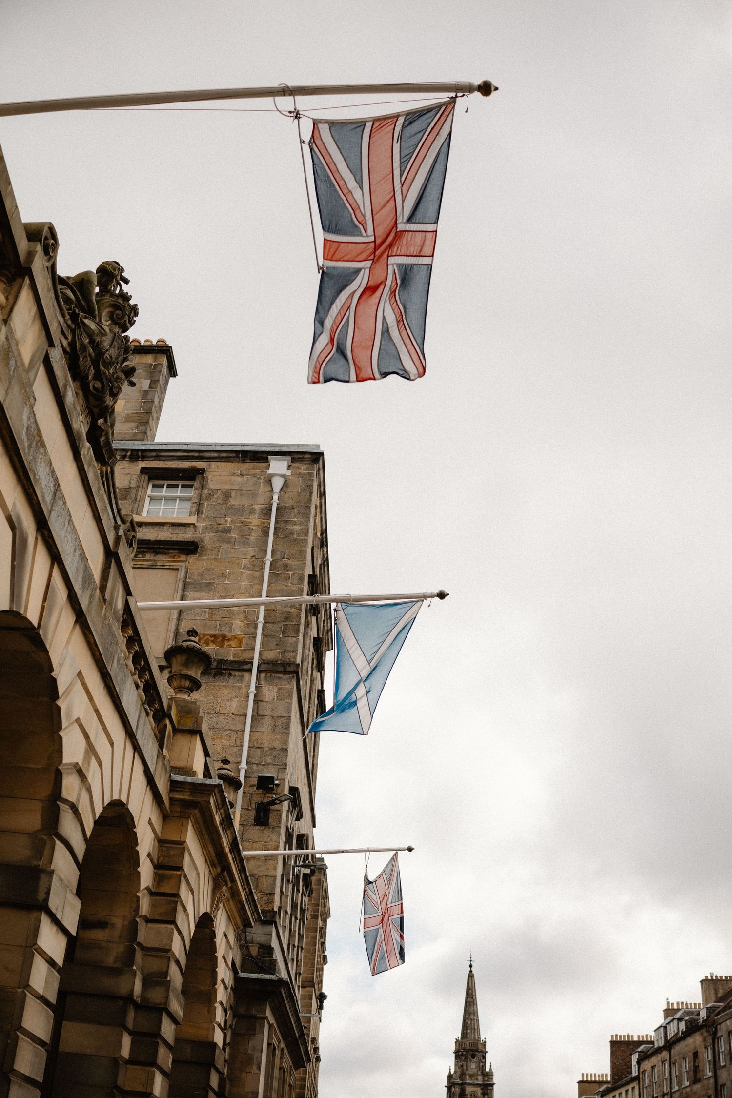Union Jack and Scottish Saltire flags hanging from historic stone building facade under overcast sky in Edinburgh.