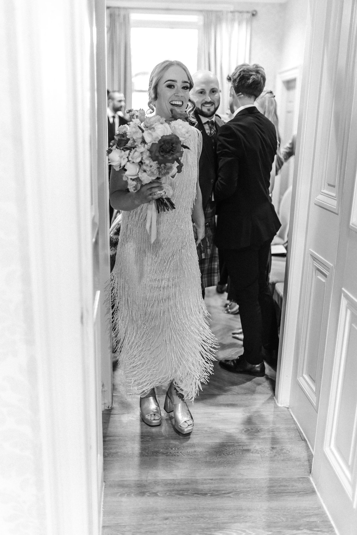 Bride in fringed white dress laughing with bouquet as groom peeks behind her through a doorway in black and white.