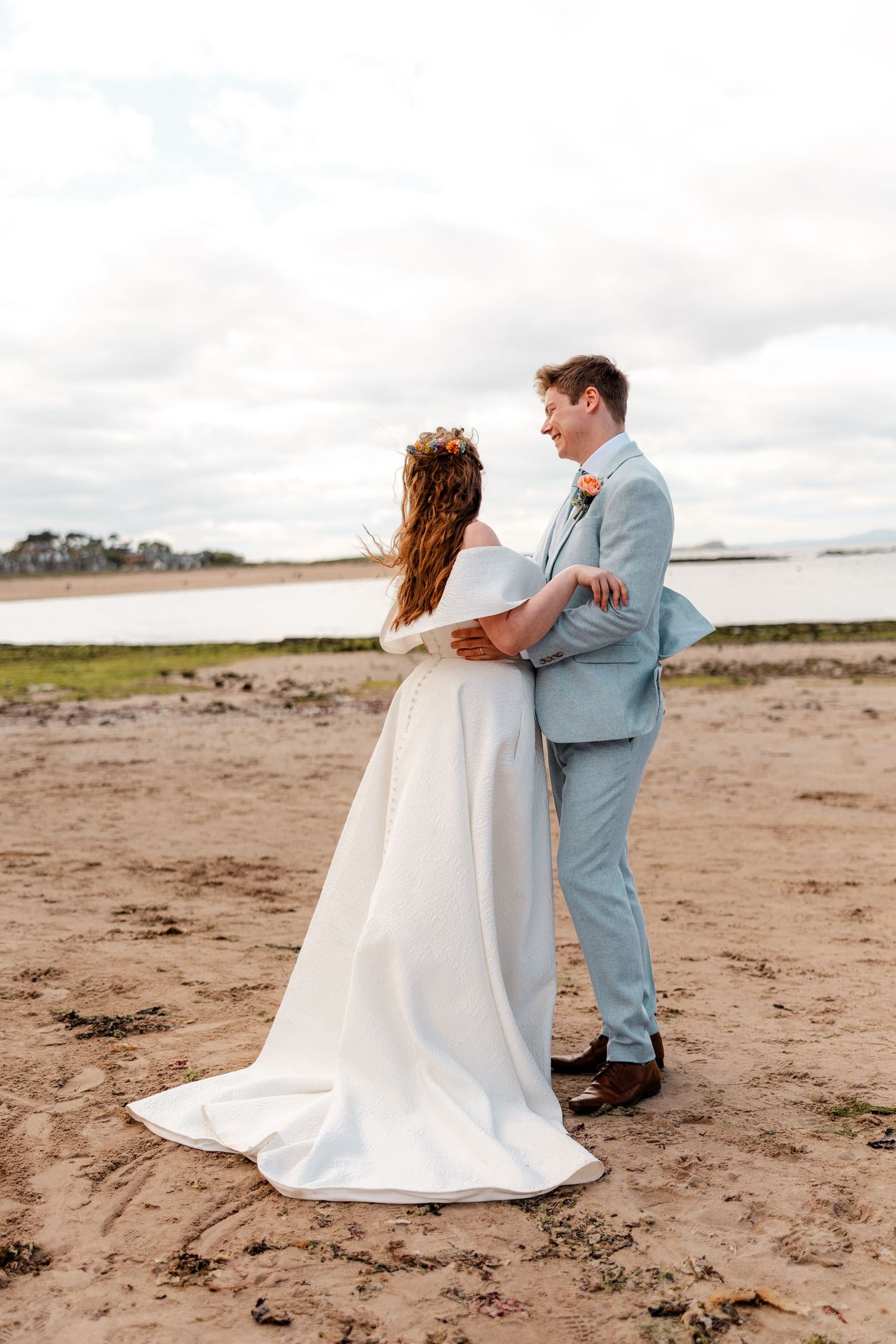 Couple in wedding attire embracing on a sandy beach under cloudy skies.