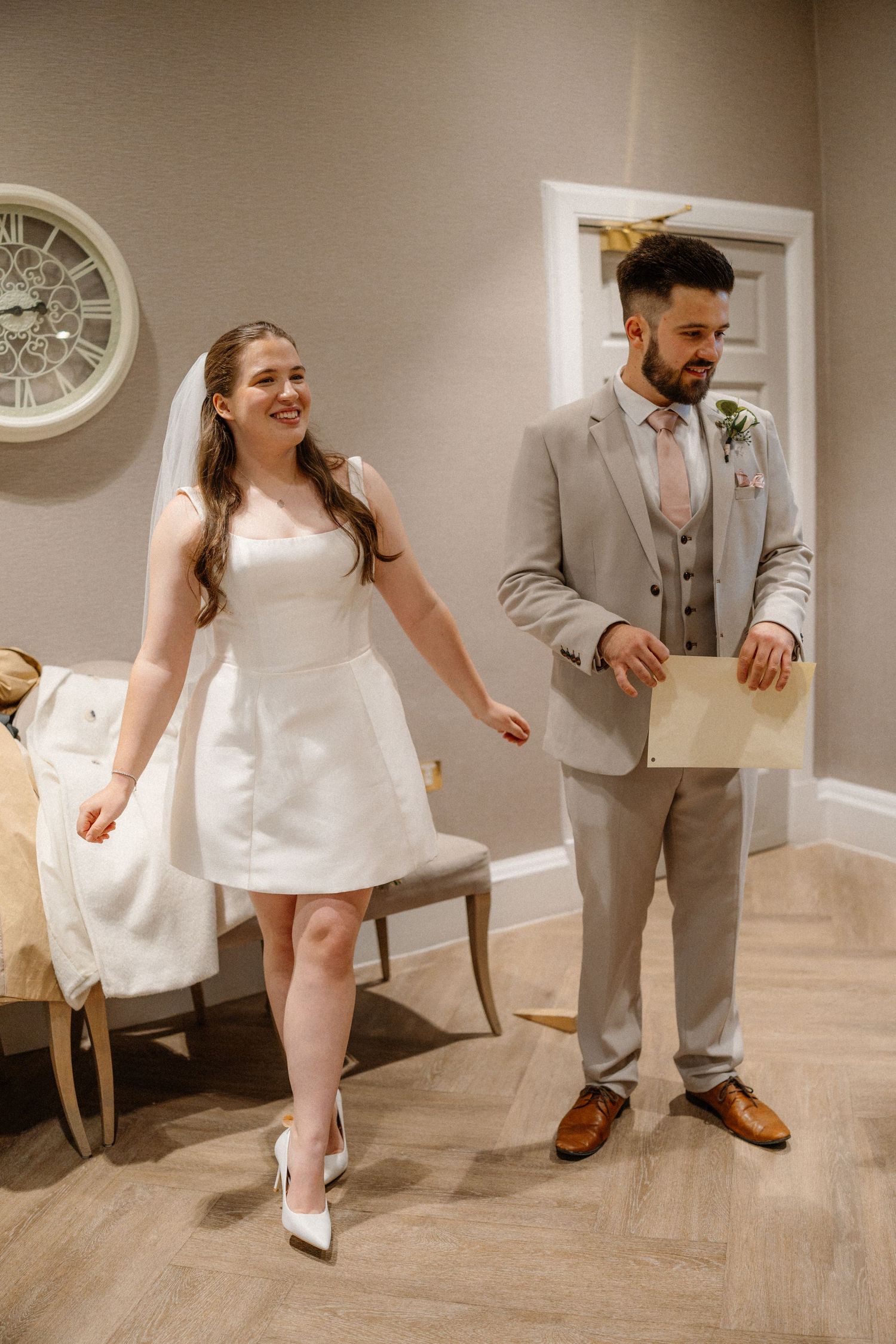 A couple in wedding attire playfully dance and pose together on a light wooden floor in a white room.