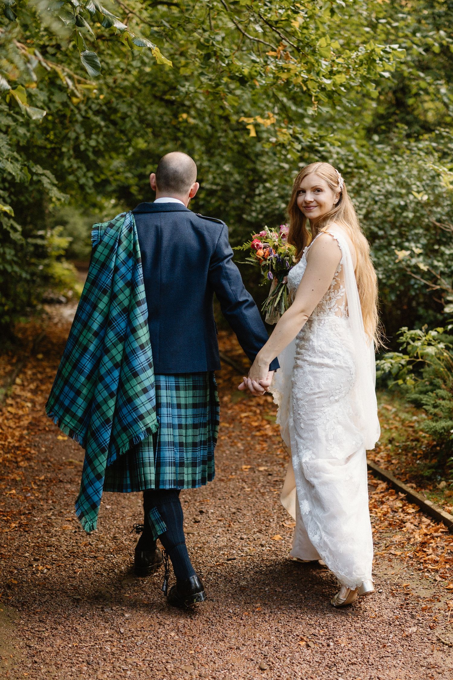 A couple walks hand in hand through an autumn forest path with the groom wearing a blue and green tartan kilt.