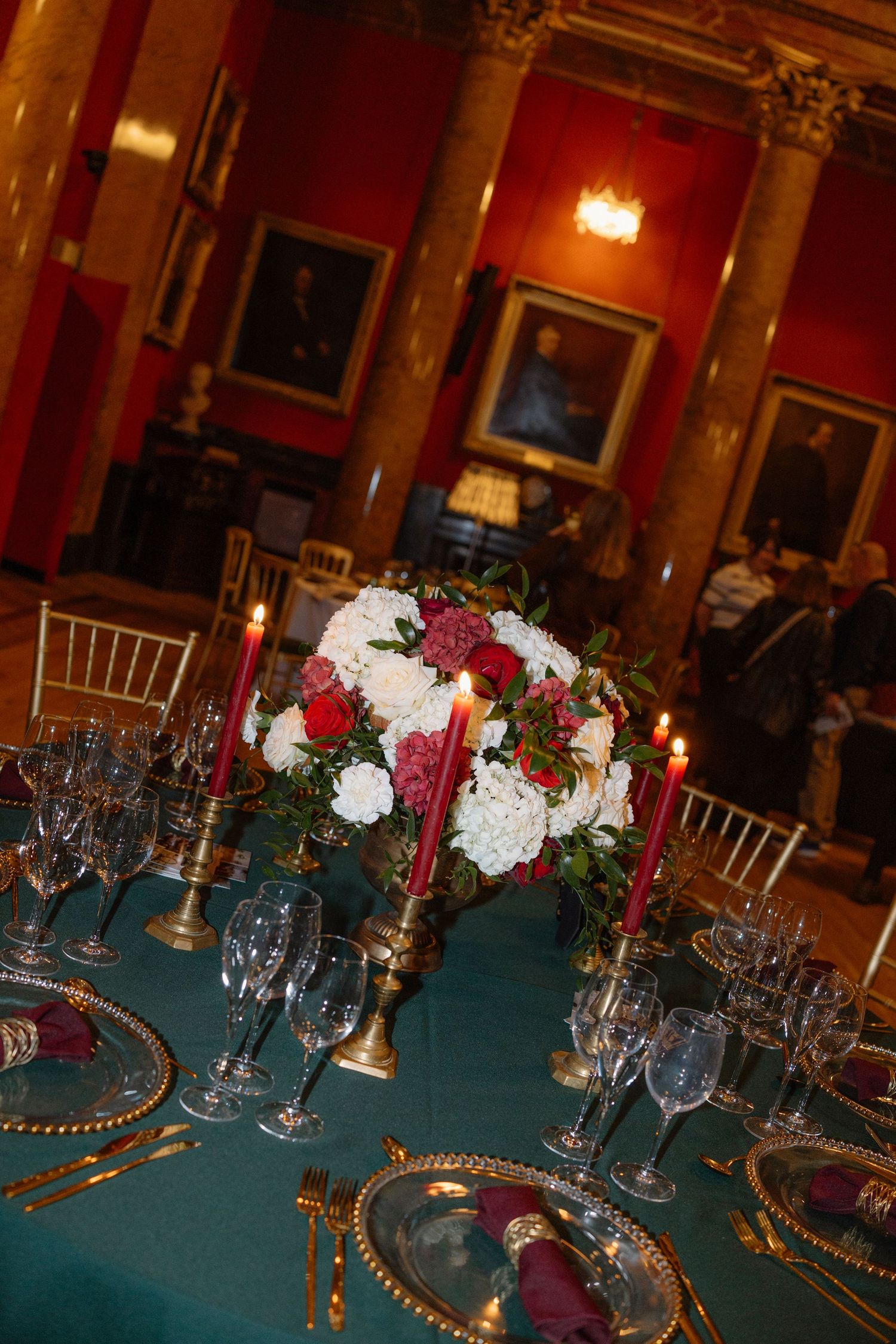 Elegant banquet table settings with burgundy and white floral centerpieces, gold chargers, and teal tablecloths.