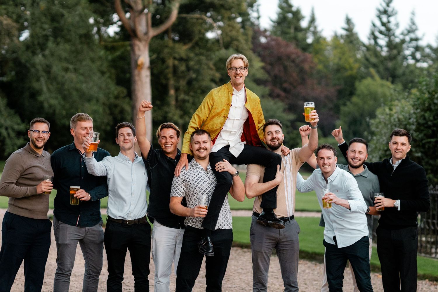 Friends celebrating outdoors with someone being lifted up against a backdrop of trees and castle architecture.