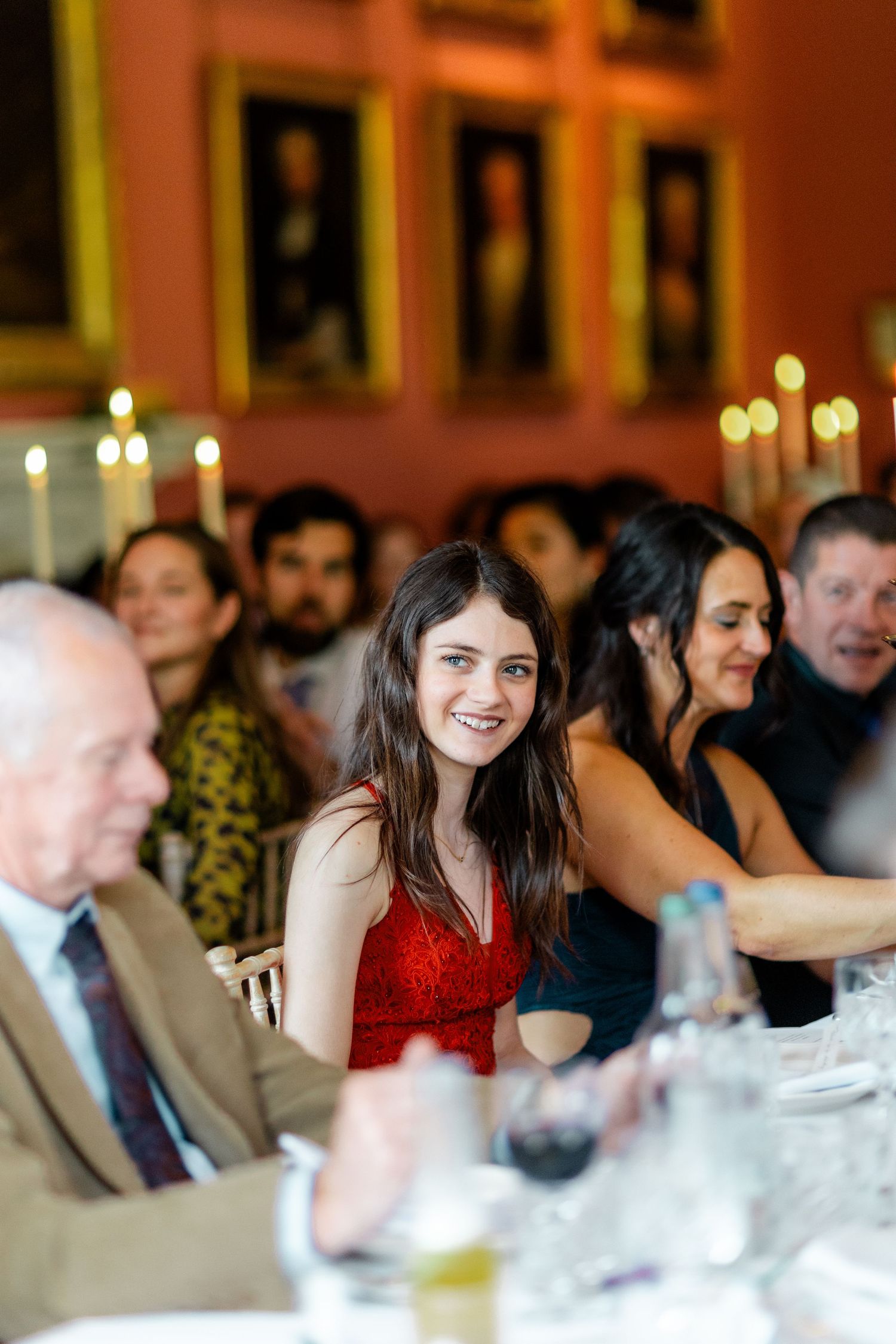 Wedding guests seated at long tables in a formal dining room with pink walls and candlelight.