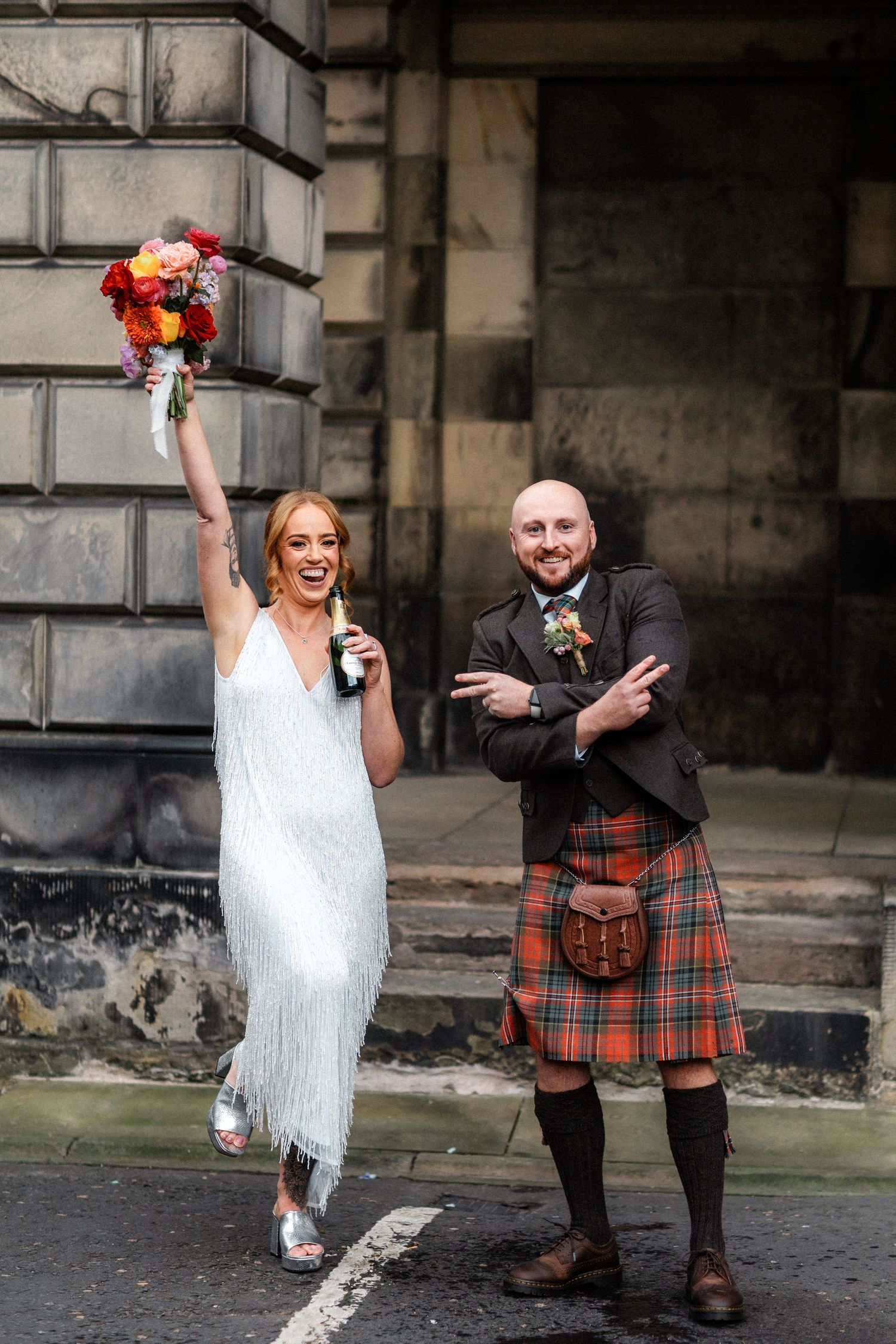 A couple poses playfully in traditional Scottish wedding attire including a kilt on the streets of Edinburgh.