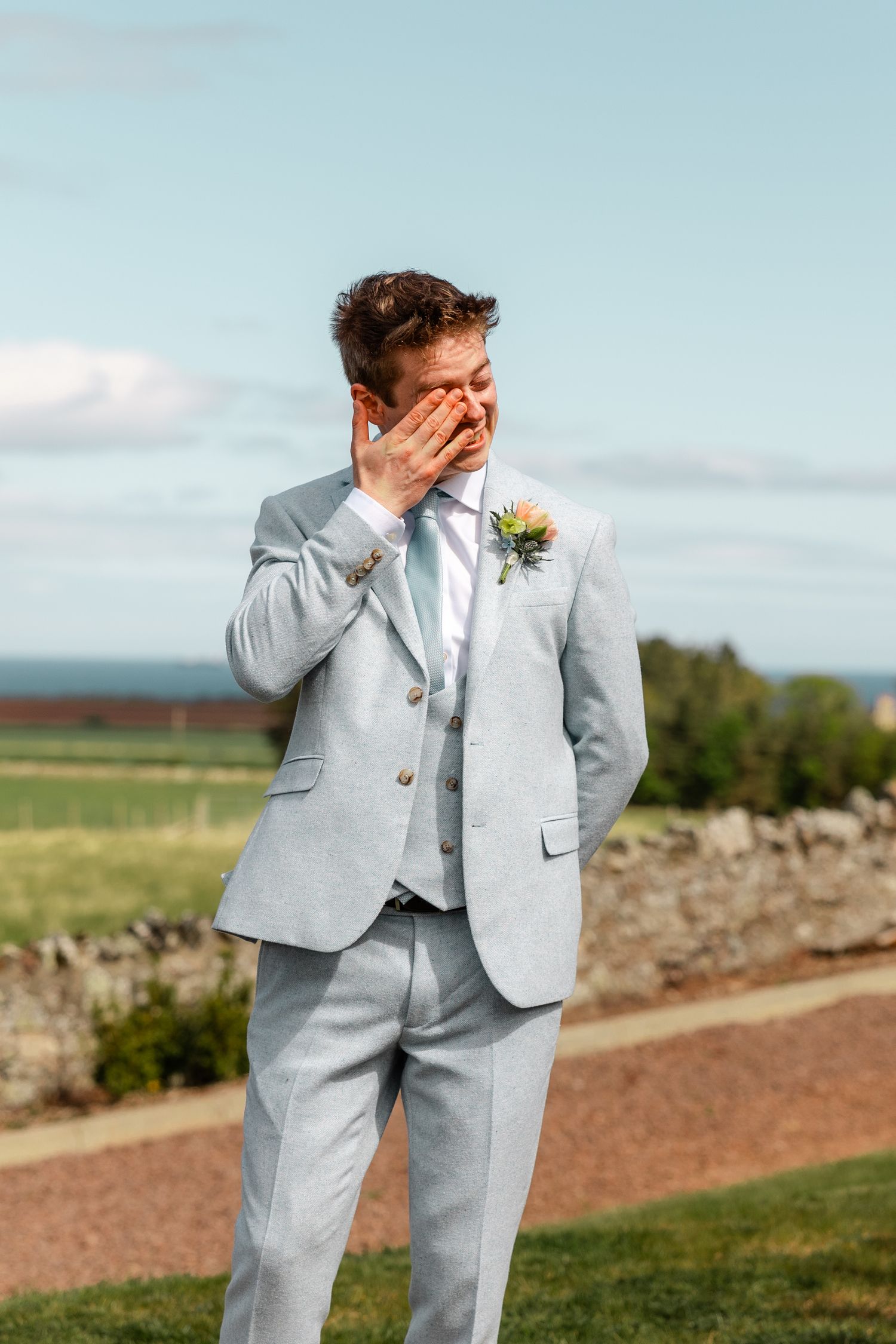 Person in light gray suit covers face with hands during emotional outdoor moment.