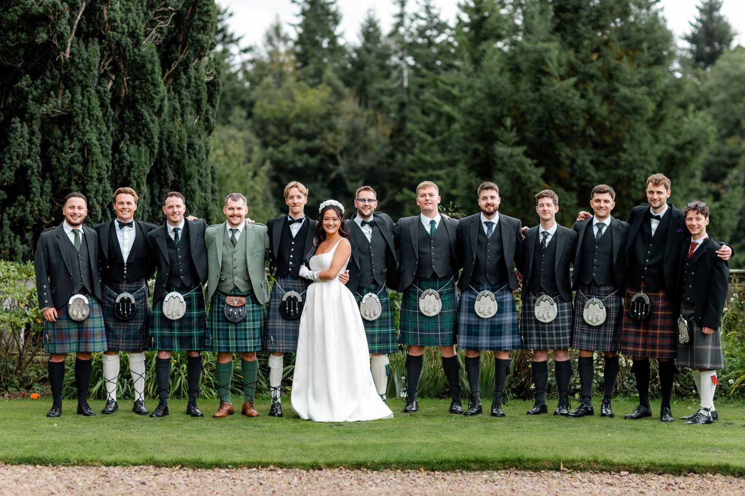 Wedding party lineup showcases mix of traditional suits and Scottish kilts outdoors.
