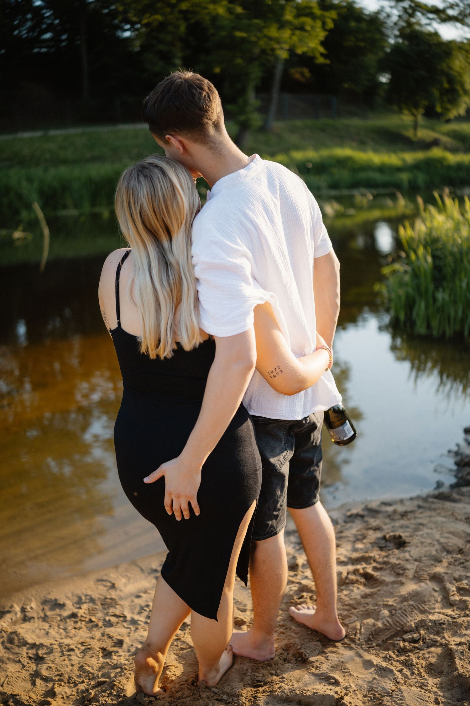 Two people walking barefoot along a lakeside beach at sunset.
