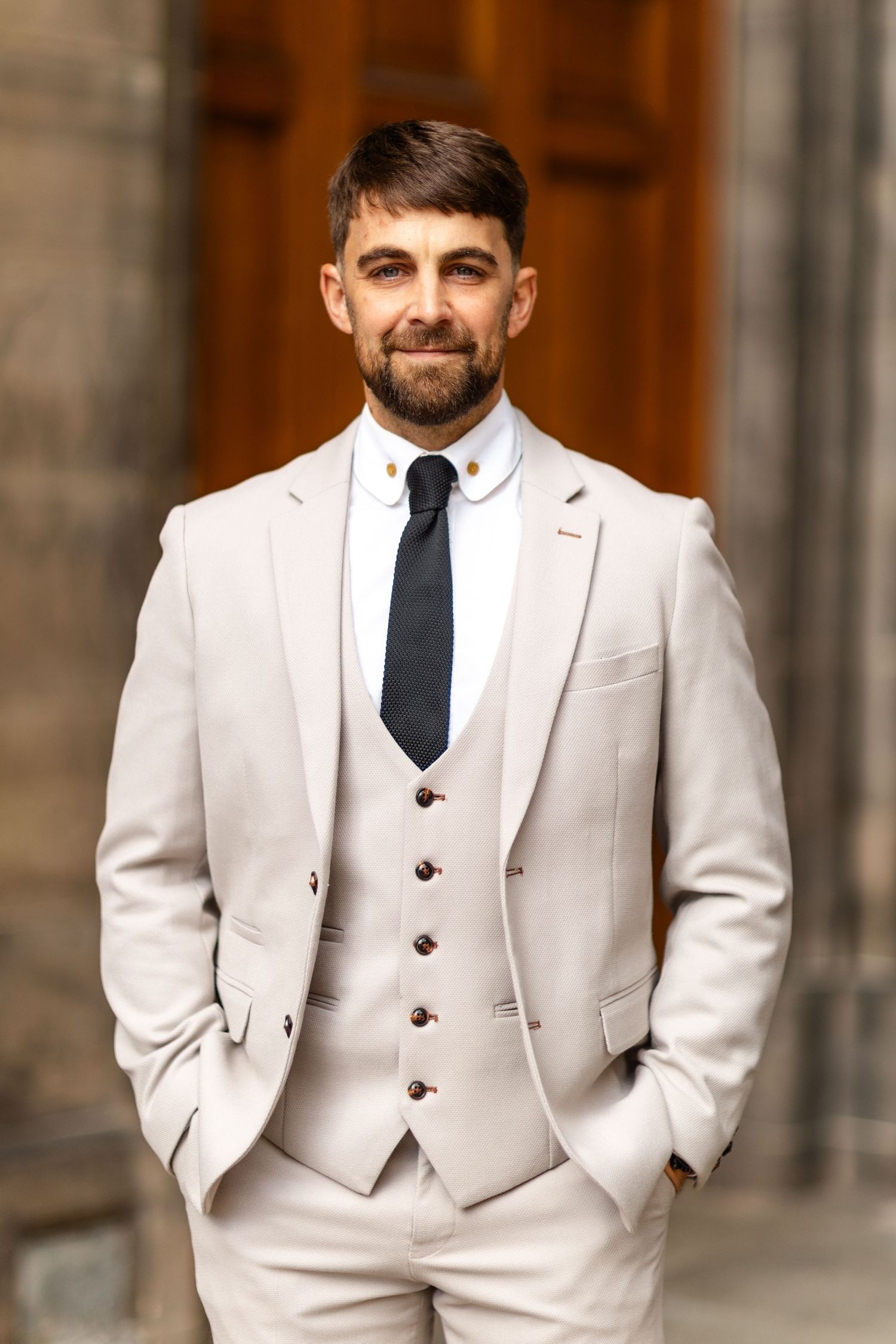 A formal portrait in a light beige three-piece suit against a classic architectural backdrop.