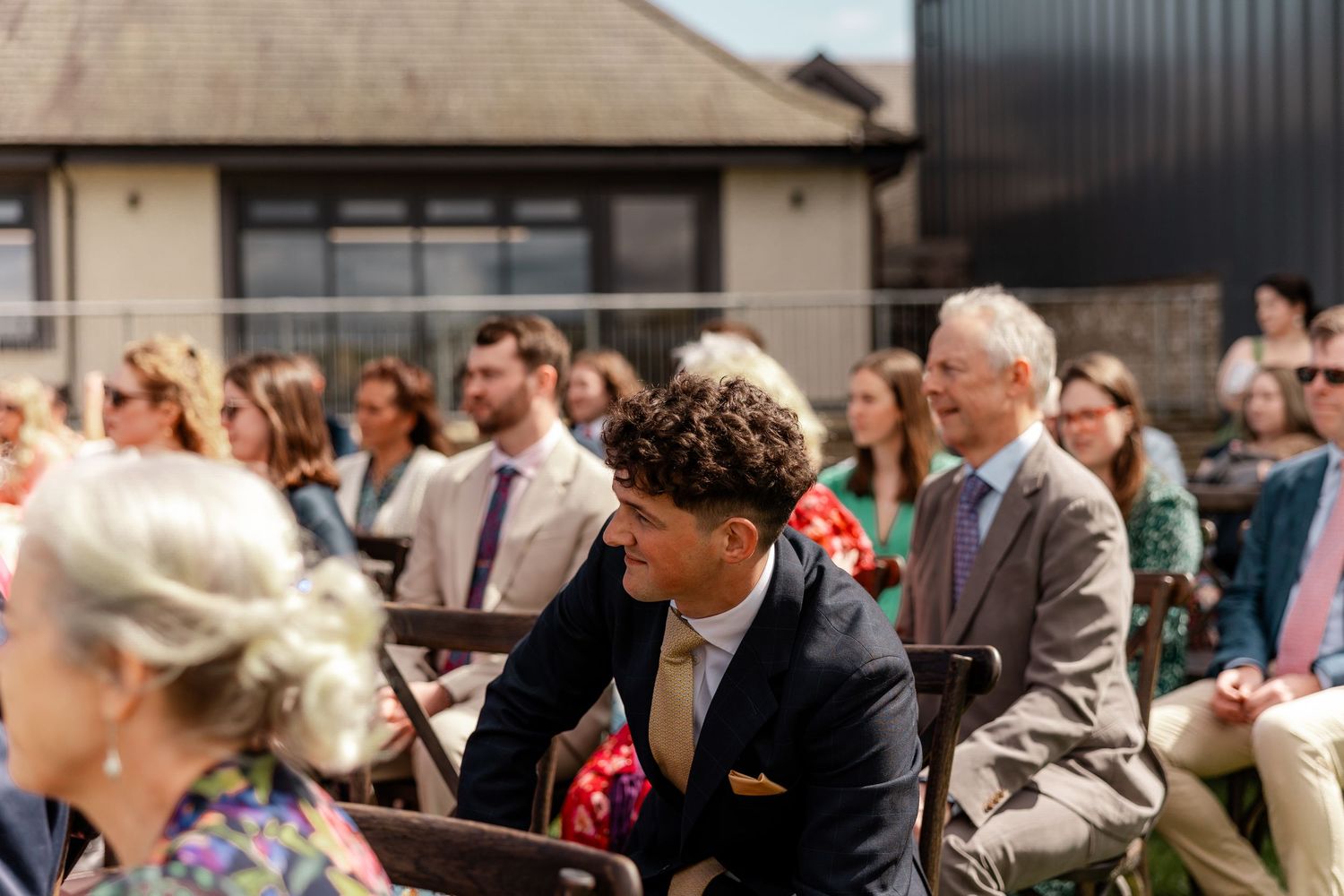 Wedding guests seated in rows outside a modern building during an outdoor ceremony.