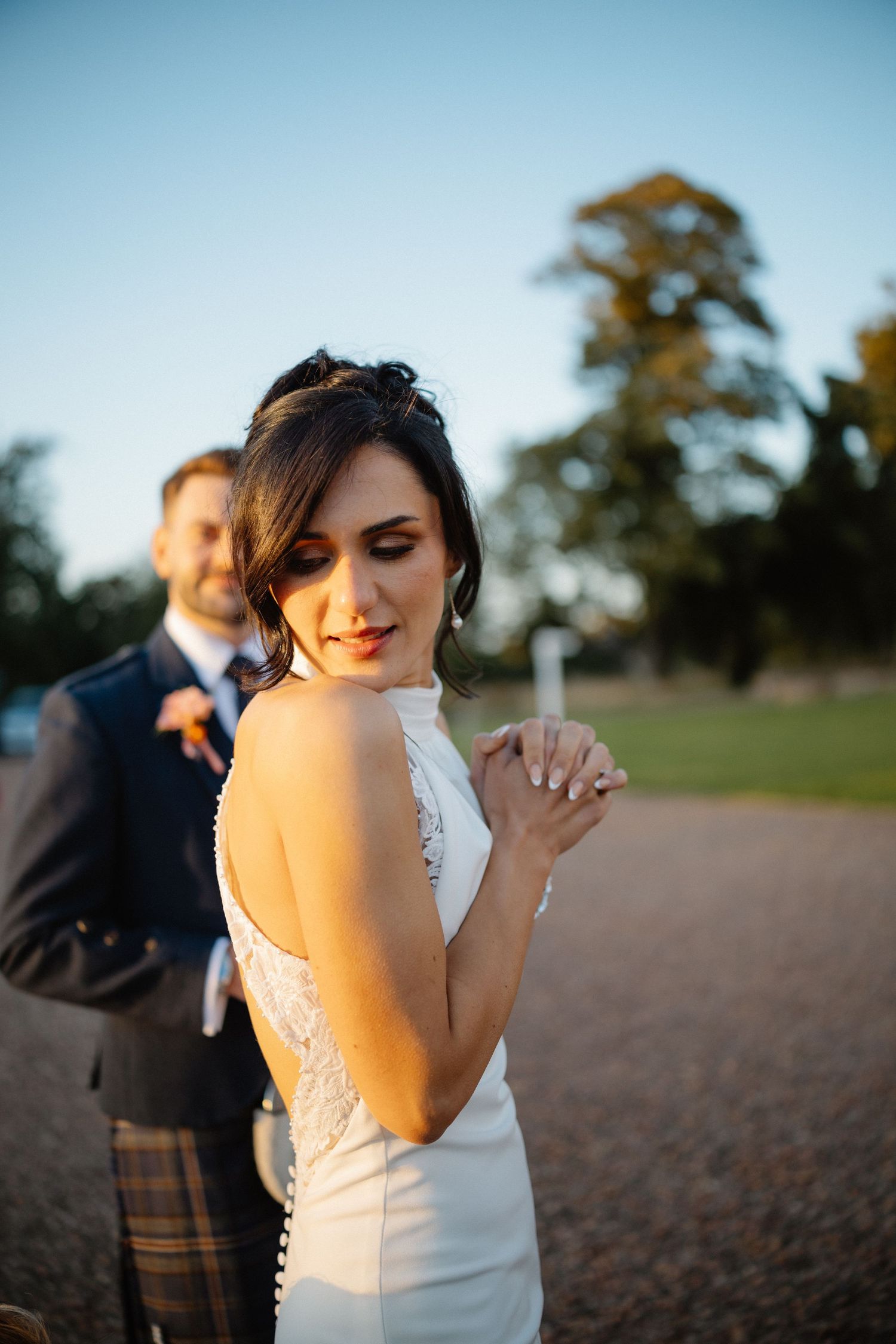 Elegant bride in white lace dress poses during golden hour outdoor photoshoot.